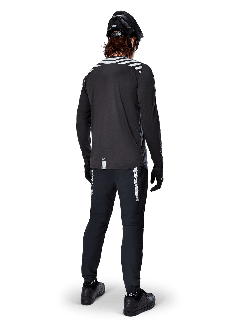 Cycliste de VTT portant le maillot à manches longues Alpinestars A-Dura Dri Oscar en coloris Black Reflective, vu de dos, associé à un pantalon technique noir, un casque et des gants, avec des graphiques à bandes réfléchissantes sur les épaules et un pan arrière allongé pour le cyclisme tout-terrain.