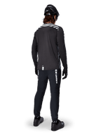 Cycliste de VTT portant le maillot à manches longues Alpinestars A-Dura Dri Oscar en coloris Black Reflective, vu de dos, associé à un pantalon technique noir, un casque et des gants, avec des graphiques à bandes réfléchissantes sur les épaules et un pan arrière allongé pour le cyclisme tout-terrain.