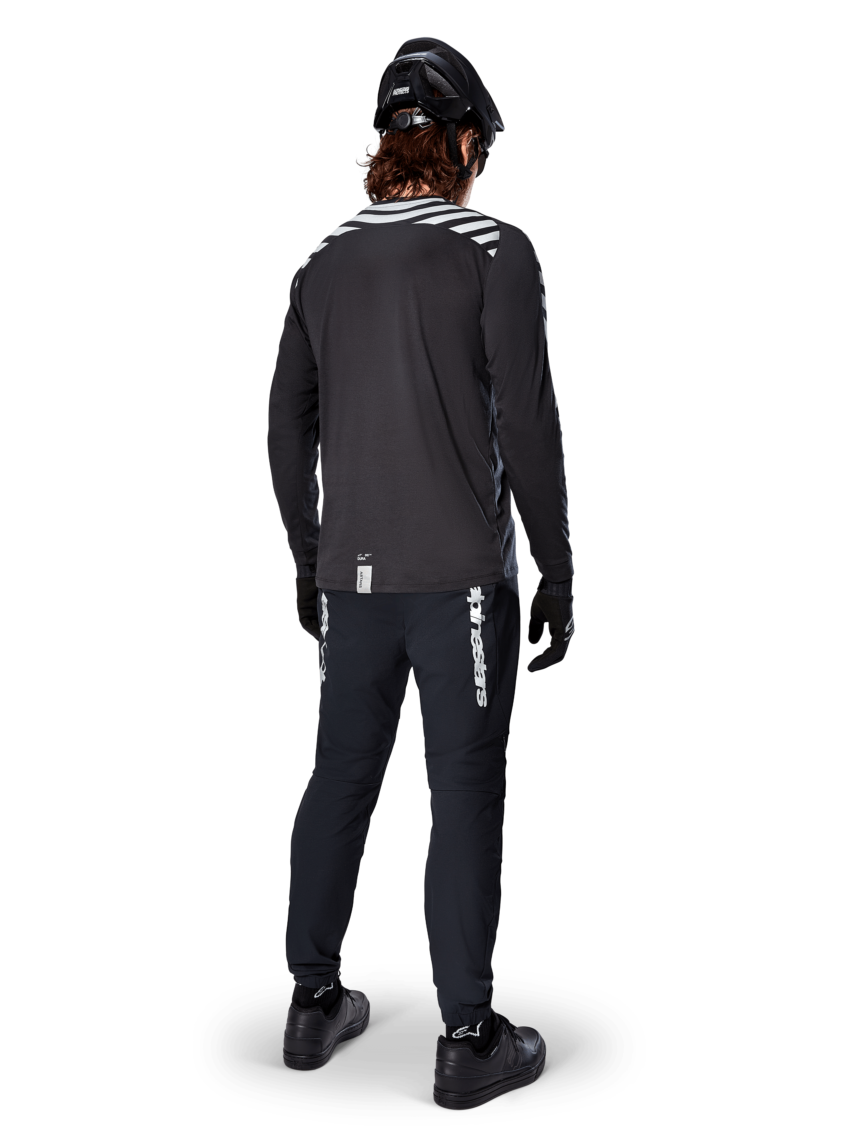 Cycliste de VTT portant le maillot à manches longues Alpinestars A-Dura Dri Oscar en coloris Black Reflective, vu de dos, associé à un pantalon technique noir, un casque et des gants, avec des graphiques à bandes réfléchissantes sur les épaules et un pan arrière allongé pour le cyclisme tout-terrain.