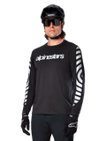 Maillot à manches longues Alpinestars A-Dura Dri Oscar, maillot de cyclisme VTT, noir réfléchissant, avec logo blanc sur la poitrine et graphismes circulaires réfléchissants sur les manches, porté avec un casque noir et des lunettes de soleil, vêtement technique évacuant l'humidité pour le VTT tout-terrain