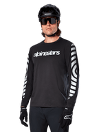 Maillot à manches longues Alpinestars A-Dura Dri Oscar, maillot de cyclisme VTT, noir réfléchissant, avec logo blanc sur la poitrine et graphismes circulaires réfléchissants sur les manches, porté avec un casque noir et des lunettes de soleil, vêtement technique évacuant l'humidité pour le VTT tout-terrain