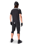 Alpinestars A-Dura Dri Oscar Short Sleeve Jersey, Black Reflective, rear view of a rider wearing the jersey with black mountain bike shorts, helmet, and knee pads, featuring reflective shoulder graphics and a drop tail design for MTB riding.