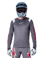 Cycliste VTT portant le maillot à manches longues Alpinestars A-Supra Astar, gris foncé avec graphismes rouges sur les manches et accents blancs aux épaules, pantalon gris assorti avec logos étoiles rouges, casque intégral bleu et masque, équipement de cyclisme tout-terrain coordonné
