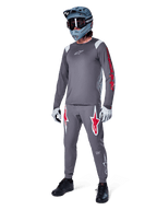 Maillot à manches longues Alpinestars A-Supra Astar gris foncé, avec marquage rouge sur les manches et logo blanc sur la poitrine, porté par un cycliste avec un pantalon gris assorti, un casque intégral bleu-gris et des gants de performance, tenue de VTT coordonnée pour la pratique de la gravité.