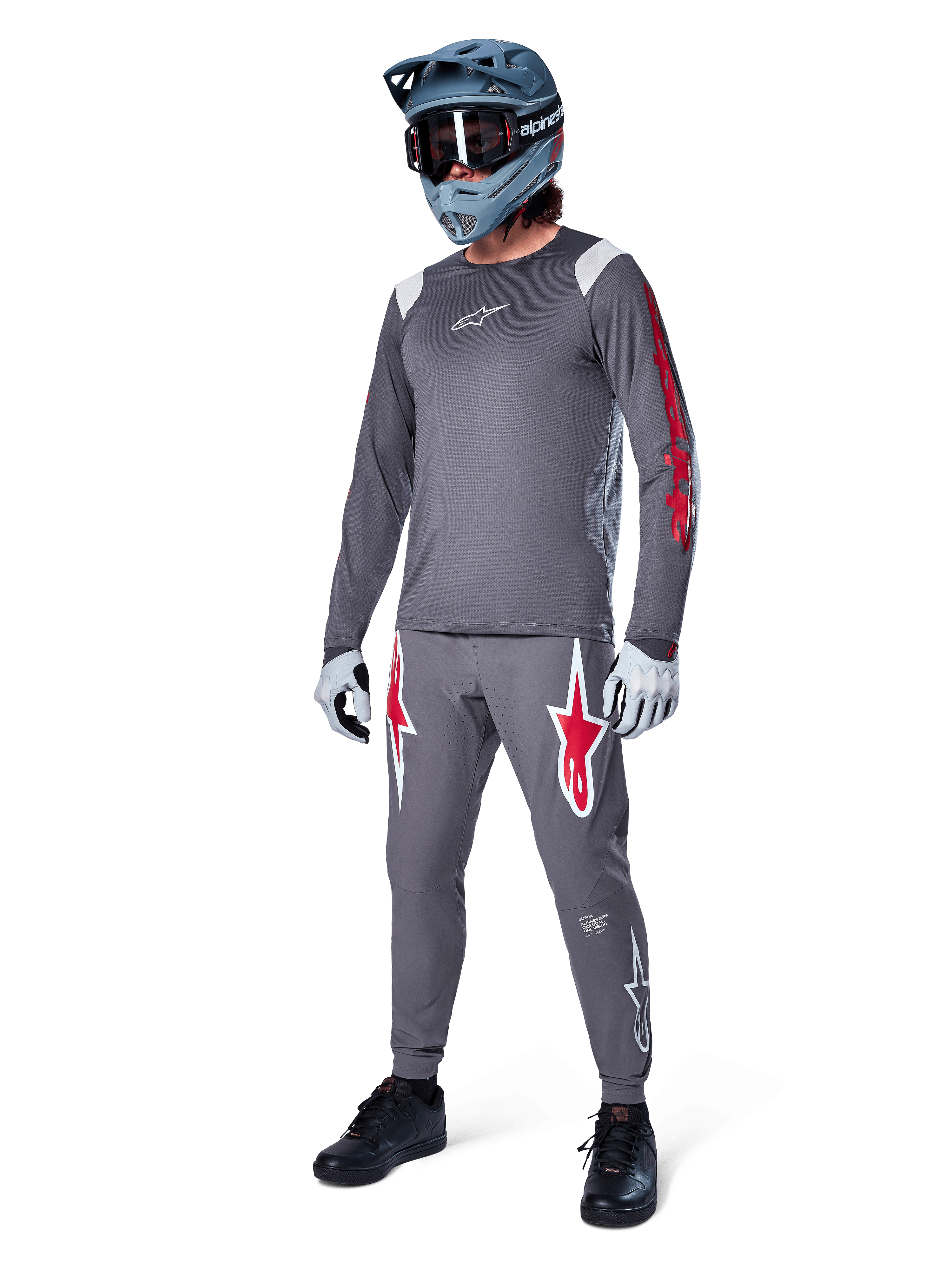 Maillot à manches longues Alpinestars A-Supra Astar gris foncé, avec marquage rouge sur les manches et logo blanc sur la poitrine, porté par un cycliste avec un pantalon gris assorti, un casque intégral bleu-gris et des gants de performance, tenue de VTT coordonnée pour la pratique de la gravité.