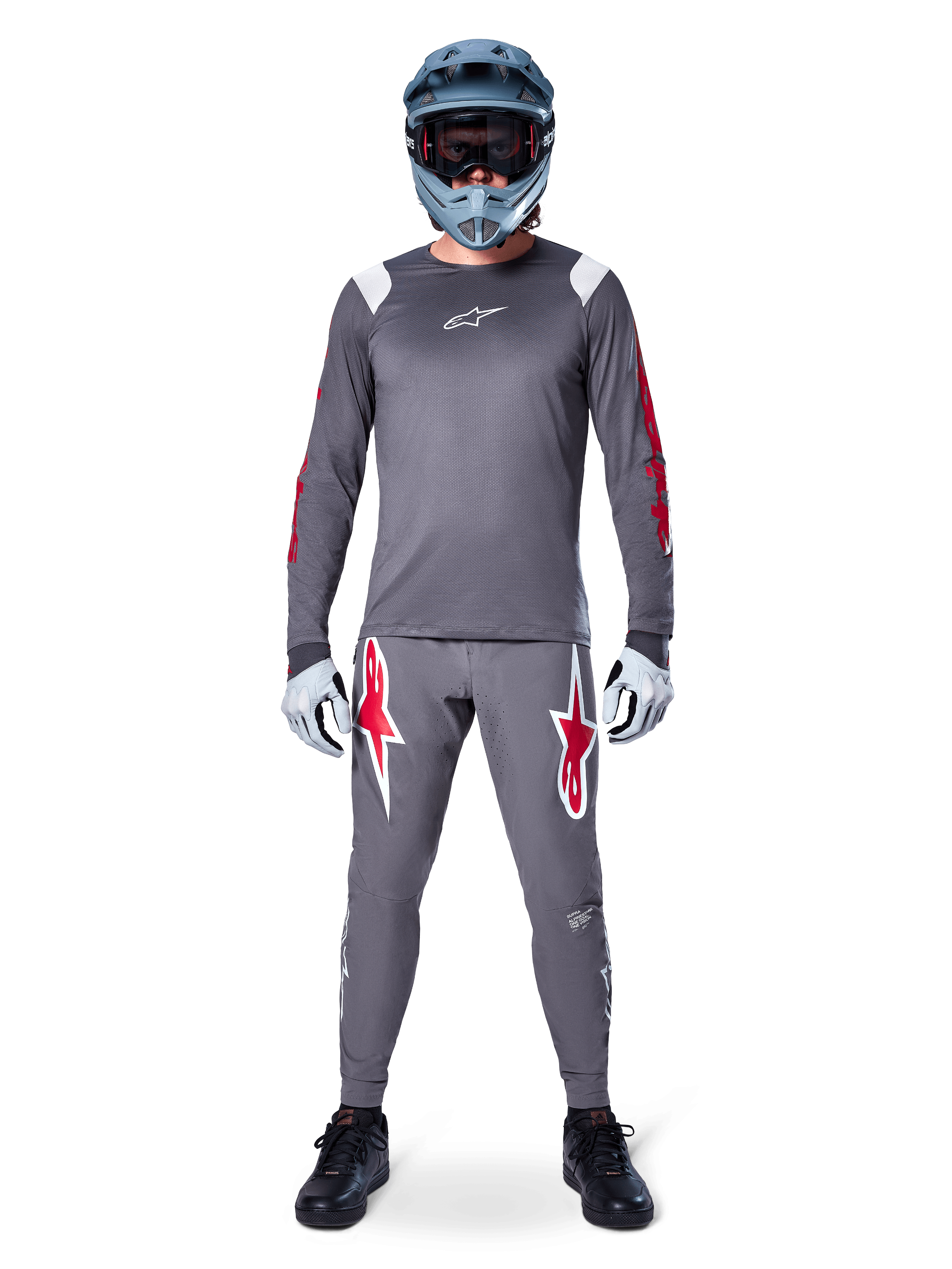 Pilote de VTT portant le maillot à manches longues Alpinestars A-Supra Astar, gris foncé avec des accents rouges et un logo blanc sur la poitrine, pantalon gris assorti avec de grands graphismes d'étoiles rouges, casque intégral bleu avec masque, gants gris clair et chaussures noires, ensemble d'équipement de VTT professionnel