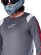 Maillot à manches longues Alpinestars A-Supra Astar, gris foncé avec empiècements blancs aux épaules et graphismes rouges sur les manches, porté avec un casque intégral bleu-gris clair, équipement de VTT haute performance