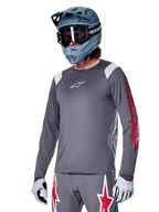 Maillot à manches longues Alpinestars A-Supra Astar, maillot de VTT, gris foncé avec des détails rouges et blancs, coupe performante, porté avec un pantalon gris assorti arborant des graphismes d'étoiles rouges et blanches, tenue de cyclisme tout-terrain complète avec casque intégral et gants