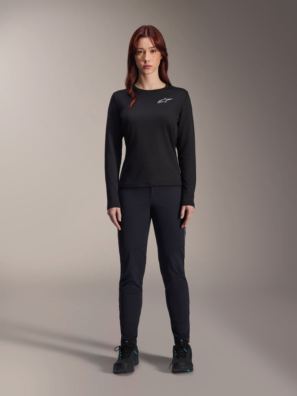 Maillot thermique Stella A-Dura d'Alpinestars, maillot de VTT à manches longues pour femme en noir, avec un logo blanc sur la poitrine, porté avec un équipement noir assorti et des chaussures de cyclisme, portrait en studio.