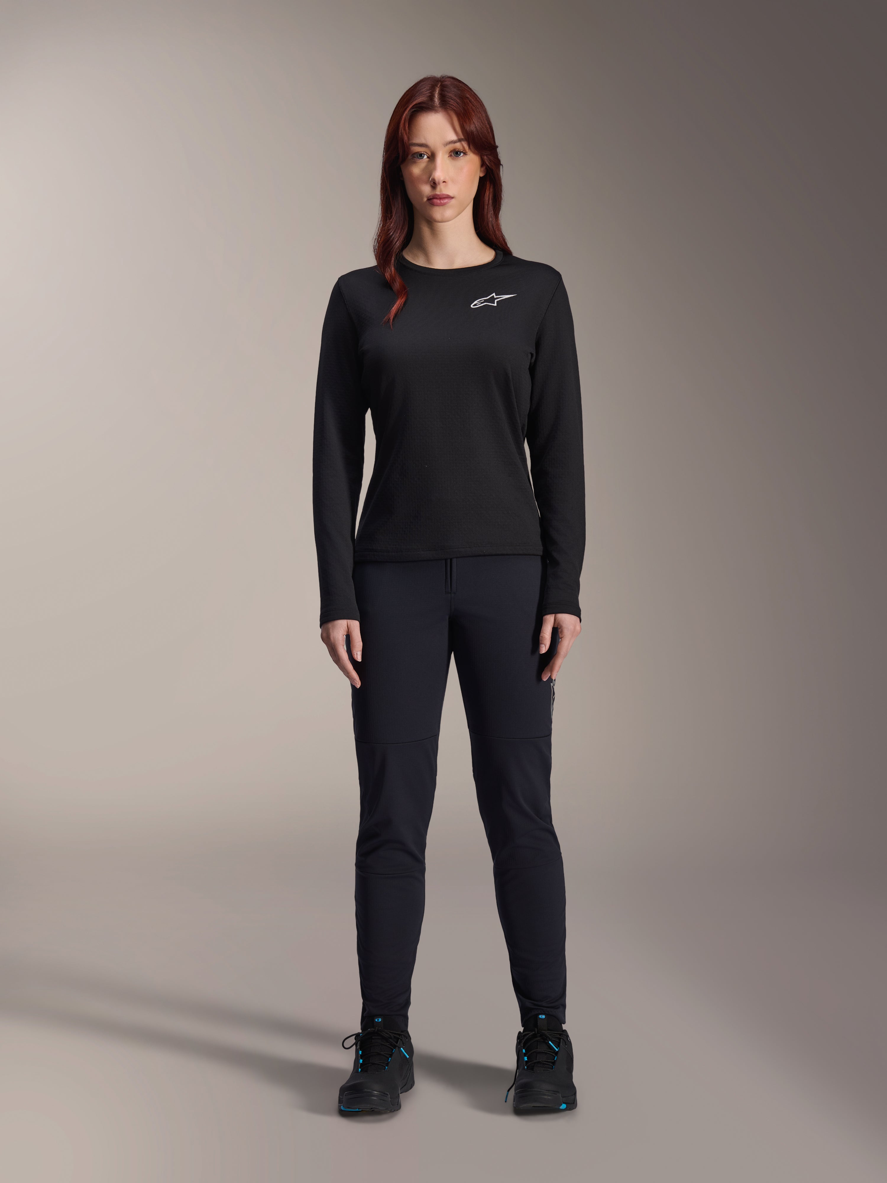 Maillot thermique Stella A-Dura d'Alpinestars, maillot de VTT à manches longues pour femme en noir, avec un logo blanc sur la poitrine, porté avec un équipement noir assorti et des chaussures de cyclisme, portrait en studio.