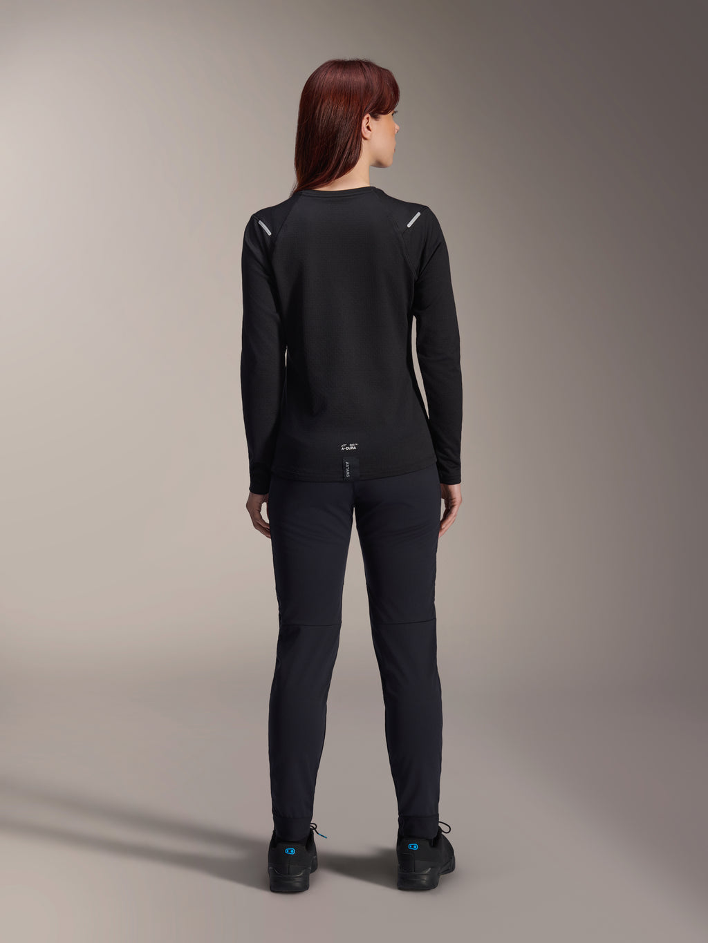 Maillot thermique Stella A-Dura Alpinestars pour femme, vue arrière, noir, maillot de cyclisme à manches longues avec polaire isolante, porté avec un pantalon et des chaussures noirs assortis, doté d'un petit marquage A-Dura en bas du dos et d'accents réfléchissants sur les épaules, conçu pour la pratique du VTT féminin.