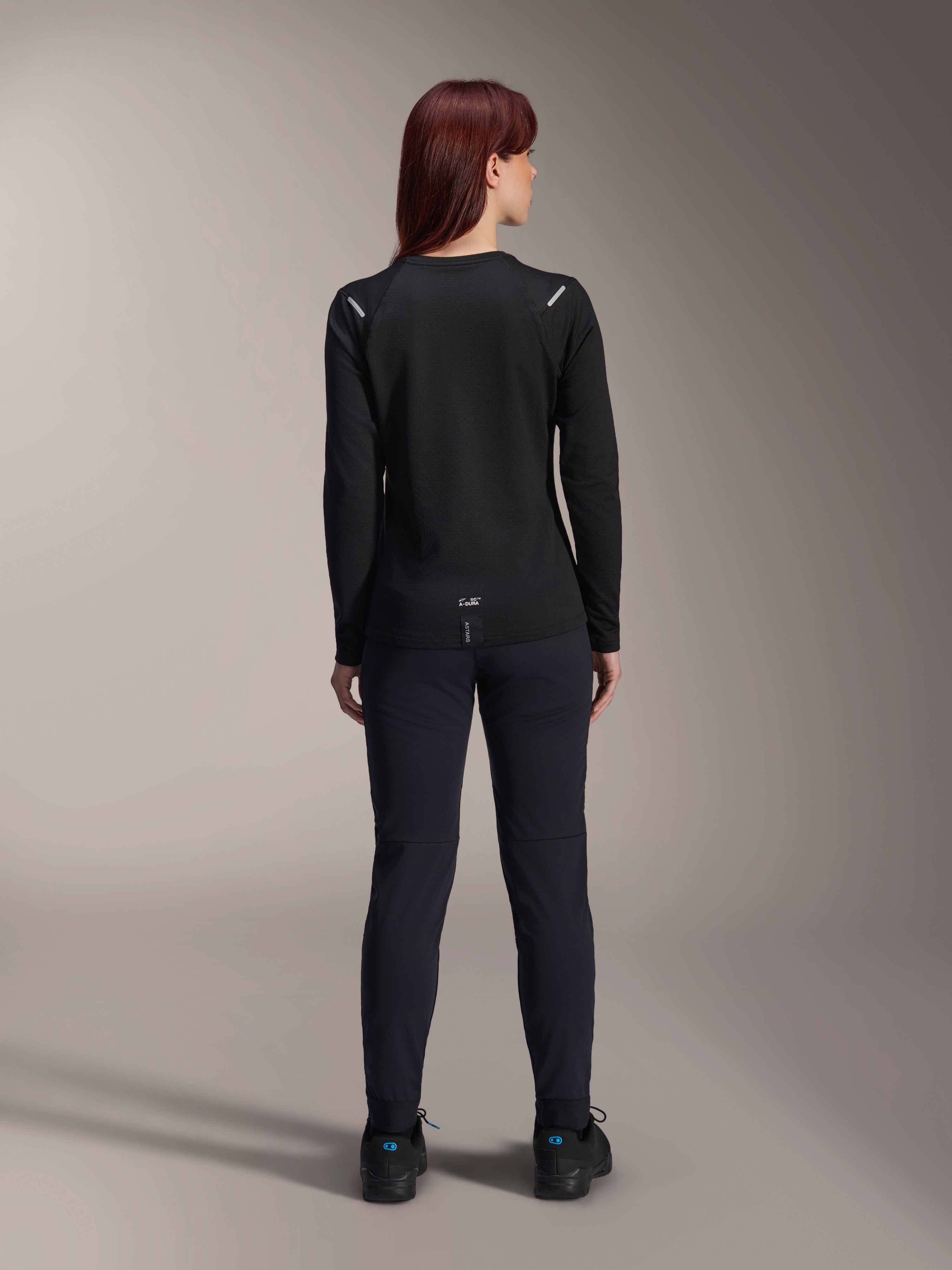 Maillot thermique Stella A-Dura Alpinestars pour femme, vue arrière, noir, maillot de cyclisme à manches longues avec polaire isolante, porté avec un pantalon et des chaussures noirs assortis, doté d'un petit marquage A-Dura en bas du dos et d'accents réfléchissants sur les épaules, conçu pour la pratique du VTT féminin.