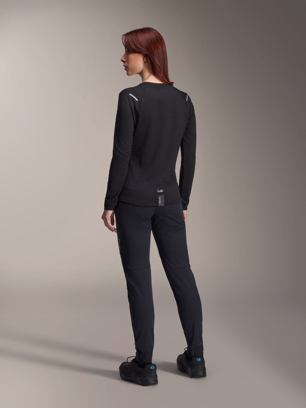 Maillot thermique pour femme Alpinestars Stella A-Dura, maillot de VTT à manches longues noir, vue arrière montrant les détails réfléchissants aux épaules et le pan arrière allongé, porté avec un pantalon et des chaussures noirs, équipement de performance pour le VTT.