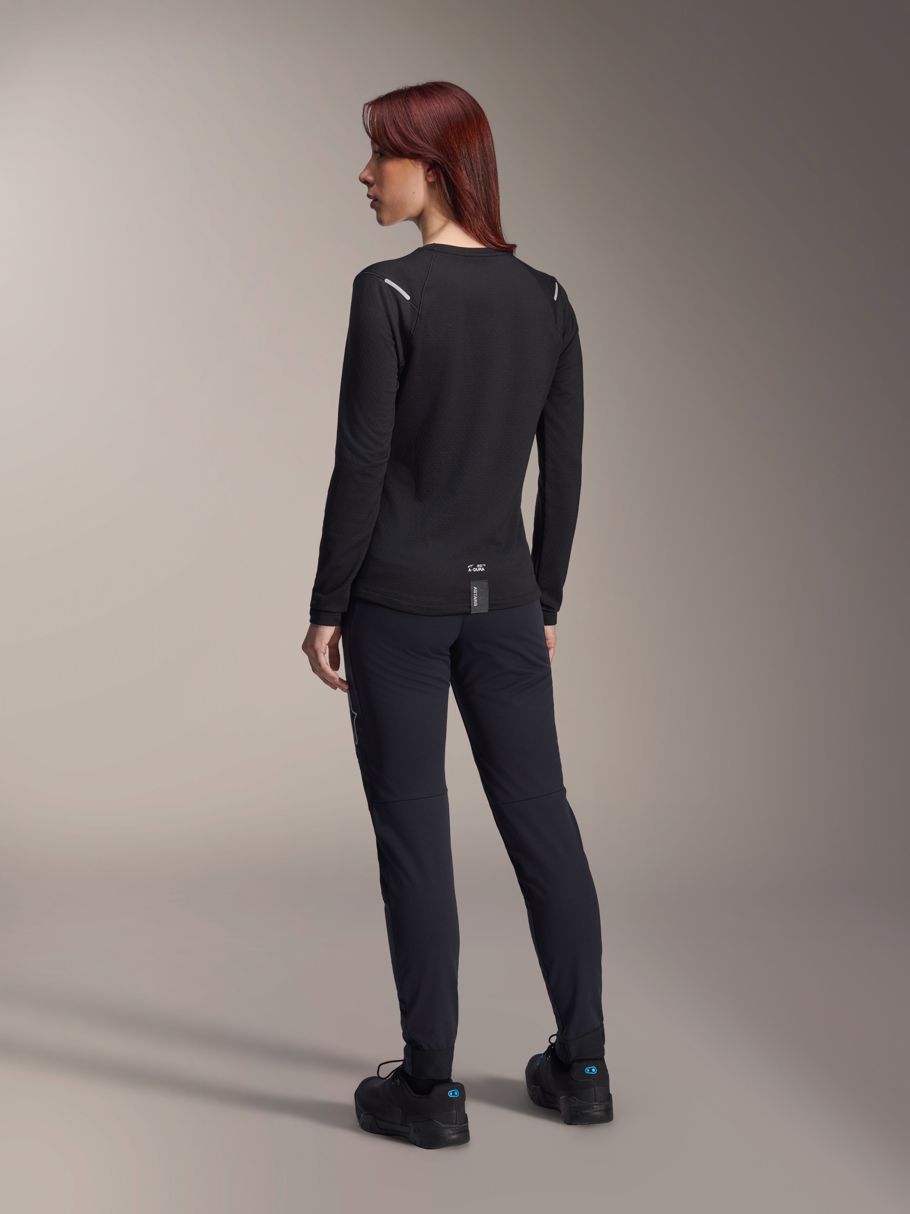 Maillot thermique pour femme Alpinestars Stella A-Dura, maillot de VTT à manches longues noir, vue arrière montrant les détails réfléchissants aux épaules et le pan arrière allongé, porté avec un pantalon et des chaussures noirs, équipement de performance pour le VTT.
