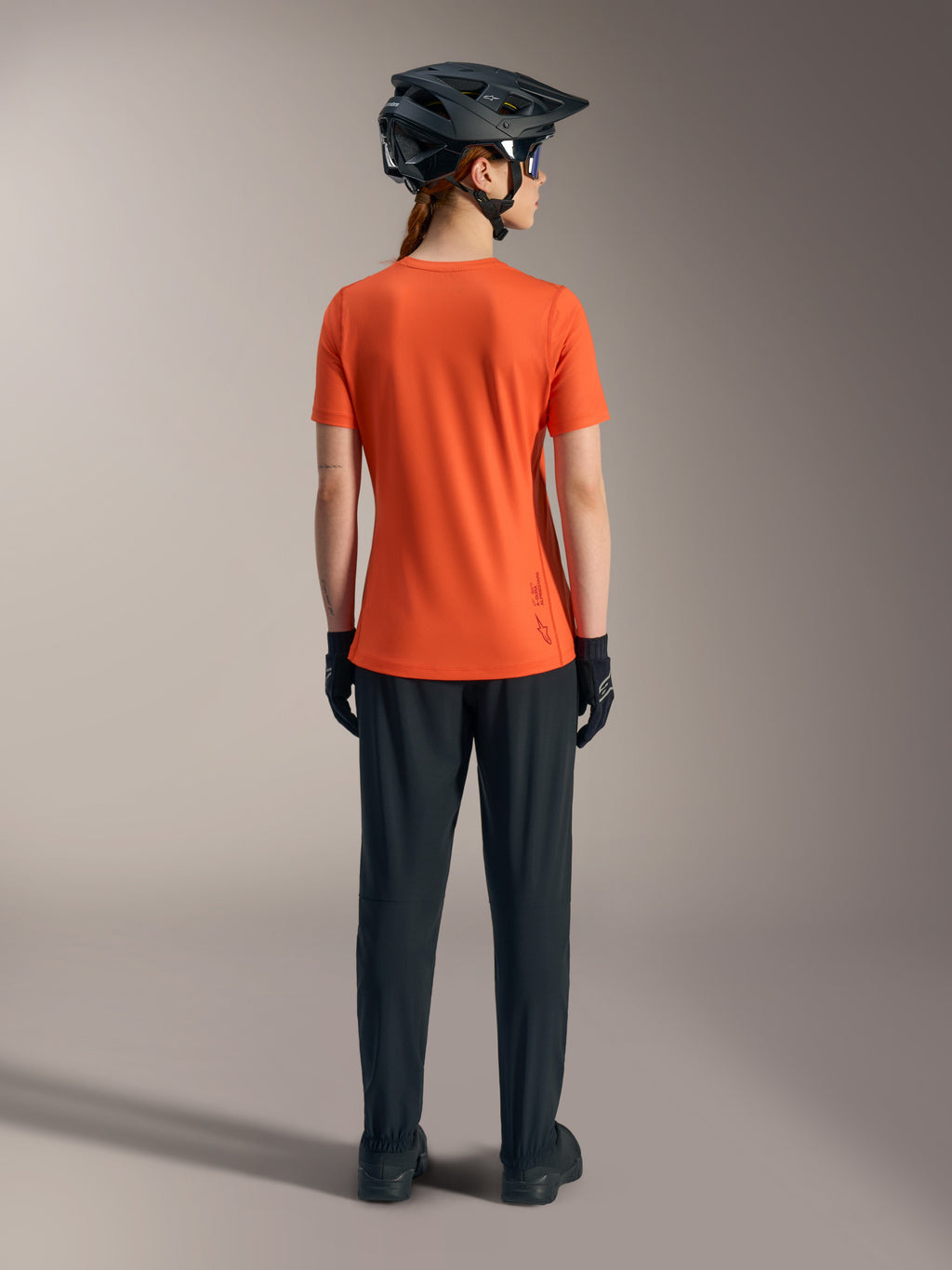 Maillot Stella A-Dura Ride Alpinestars orange pour femme, vue de dos avec pantalon de VTT noir et casque.