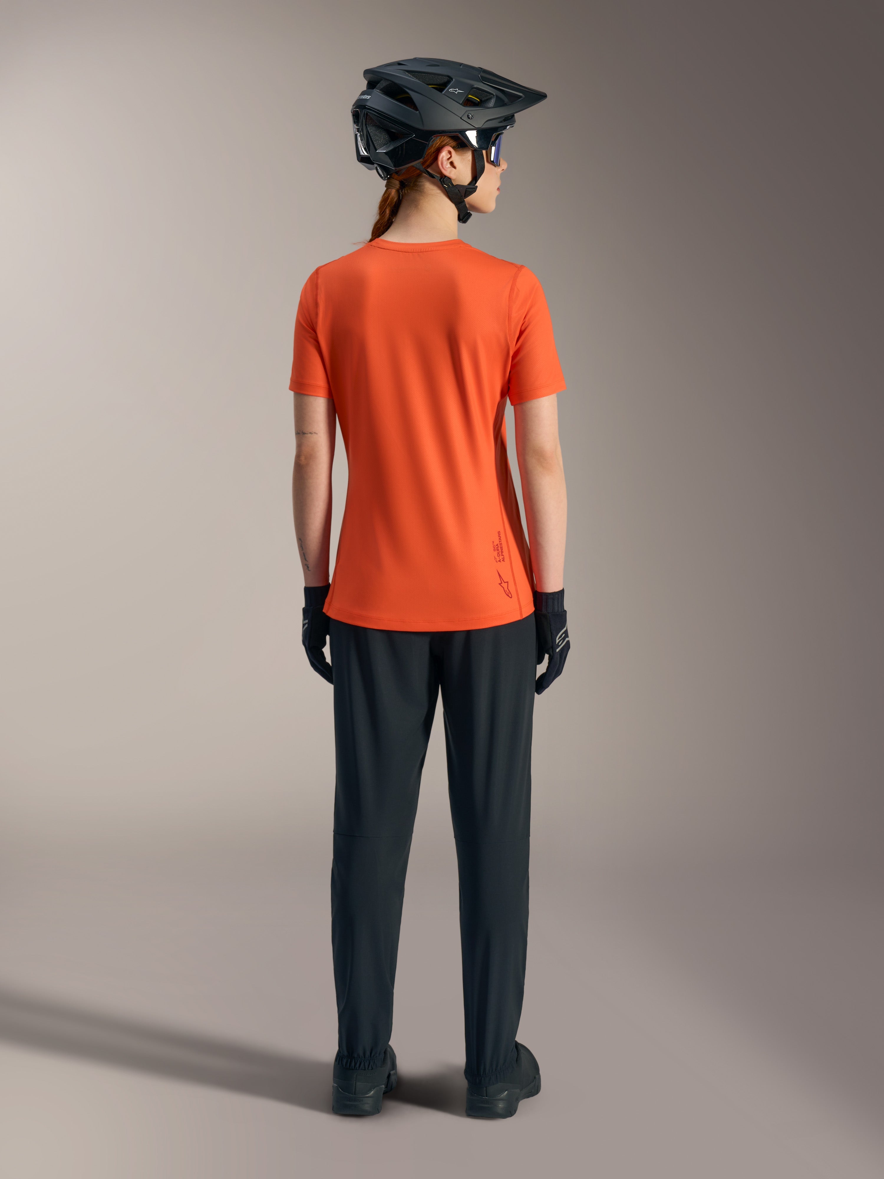 Maillot Stella A-Dura Ride Alpinestars orange pour femme, vue de dos avec pantalon de VTT noir et casque.