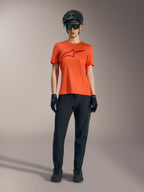 Maillot femme Stella A-Dura Ride orange à manches courtes, logo bordeaux, avec équipement VTT, casque et gants noirs.