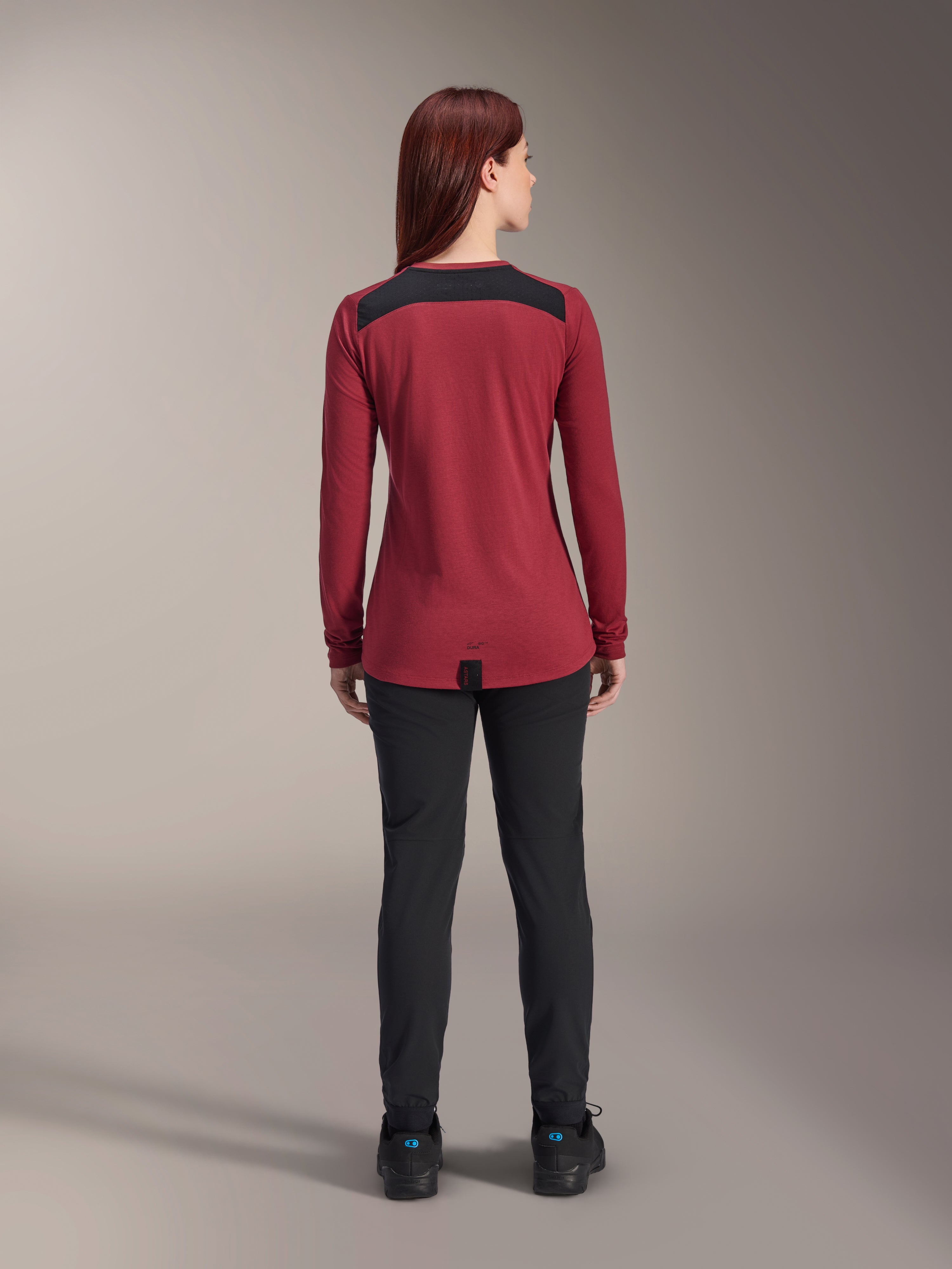 Maillot à manches longues Alpinestars Stella A-Dura Dri Astar pour femme, rouge bordeaux avec empiècements en maille noire sur les épaules, vêtement technique de VTT vu de dos avec pantalon noir, design à pan arrière allongé pour une protection optimale à vélo