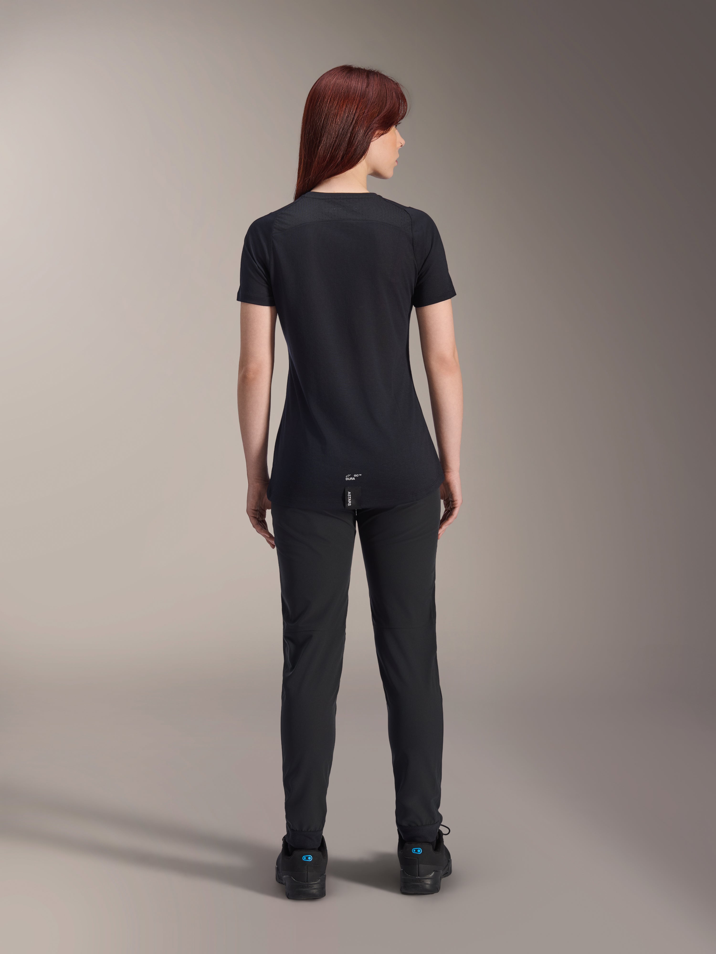Maillot à manches courtes Alpinestars Stella A-Dura Dri Switch pour femme, noir, vue arrière sur un modèle, avec un pan arrière allongé et des panneaux en maille technique, associé à un pantalon noir, équipement de cyclisme VTT de performance