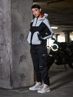 Pantalon Actea Femme Alpinestars, noir, motarde portant un pantalon de moto de style cargo avec une veste technique grise et noire et des baskets beiges, cadre de parking urbain avec une moto.