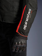 Veste Alpinestars T-Sps Air V2, gros plan sur la manche en Noir Rouge Fluo, logos Alpinestars et T-SPS AIR blancs, bande rouge fluorescent, portée avec un gant de moto protecteur noir avec protection des articulations en fibre de carbone
