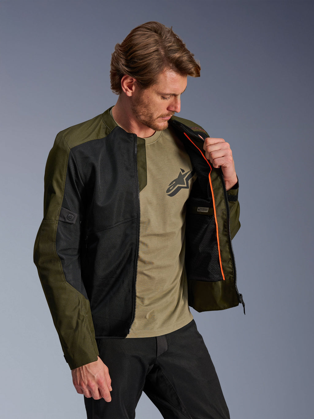 Veste Alpinestars C-1 Air en kaki et noir, portée par un mannequin homme pour montrer la doublure intérieure orange et le compartiment imperméable, dotée de grands panneaux en maille technique pour la circulation de l'air, style moto urbain contemporain associé à un t-shirt olive et un pantalon noir