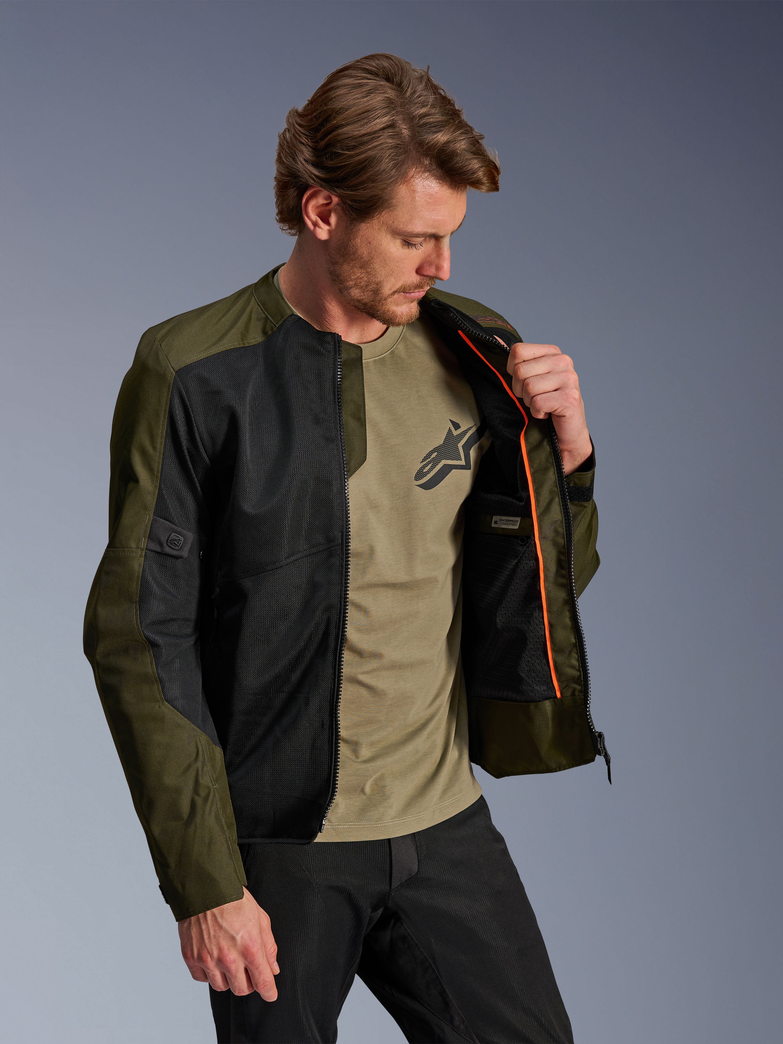 Veste Alpinestars C-1 Air en kaki et noir, portée par un mannequin homme pour montrer la doublure intérieure orange et le compartiment imperméable, dotée de grands panneaux en maille technique pour la circulation de l'air, style moto urbain contemporain associé à un t-shirt olive et un pantalon noir