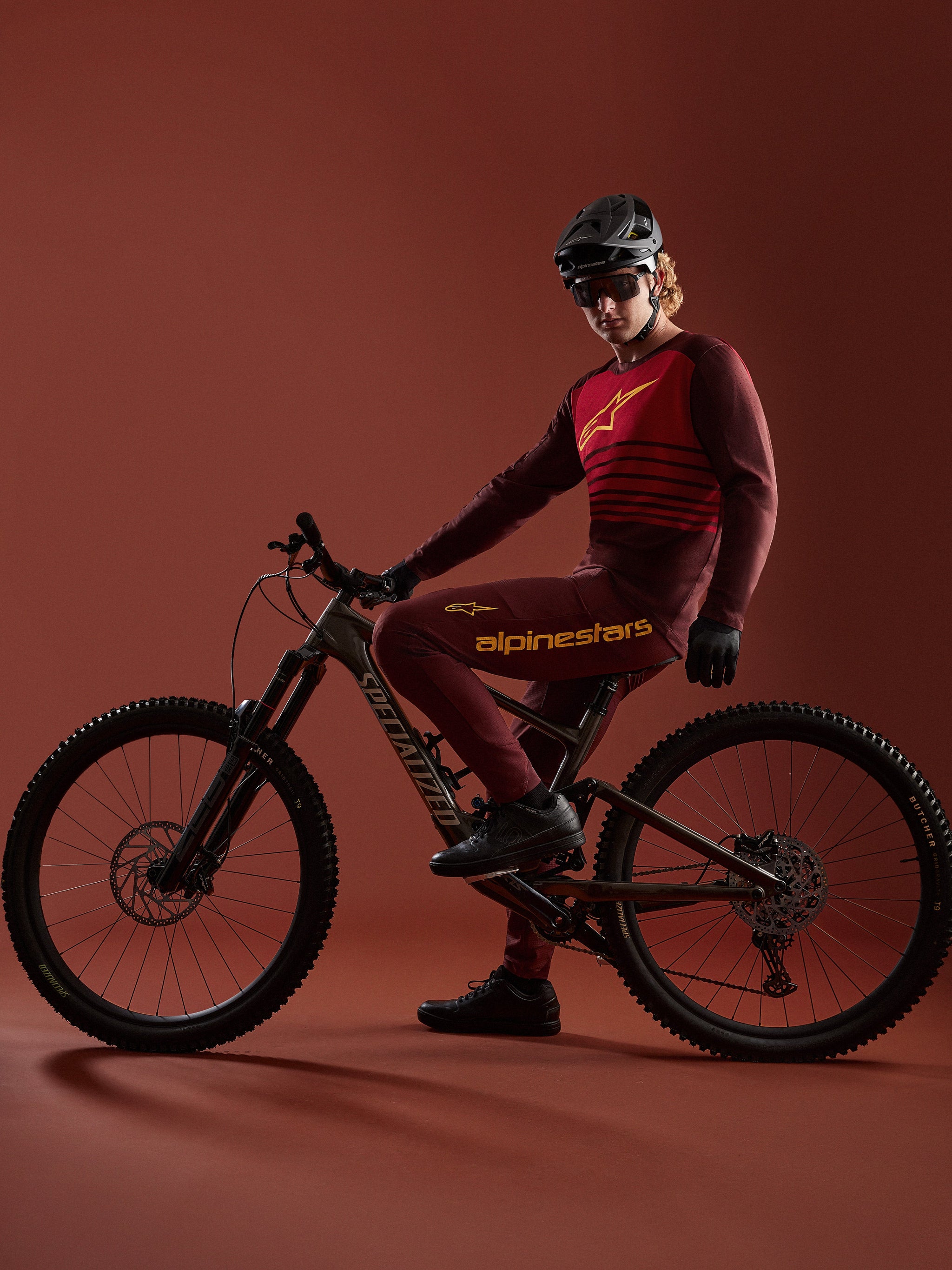 Maillot bordeaux A-Dura Elite 2.0 et pantalon assorti Alpinestars portés par un vététiste sur vélo Specialized en studio.