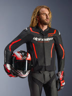 Alpinestars GP Force V2 Leather Jacket, Black and Red Fluo, worn by a male model holding a matching helmet, featuring white Alpinestars logos and red accents, paired with leather pants and racing gloves, sport motorcycle gear for road and track.