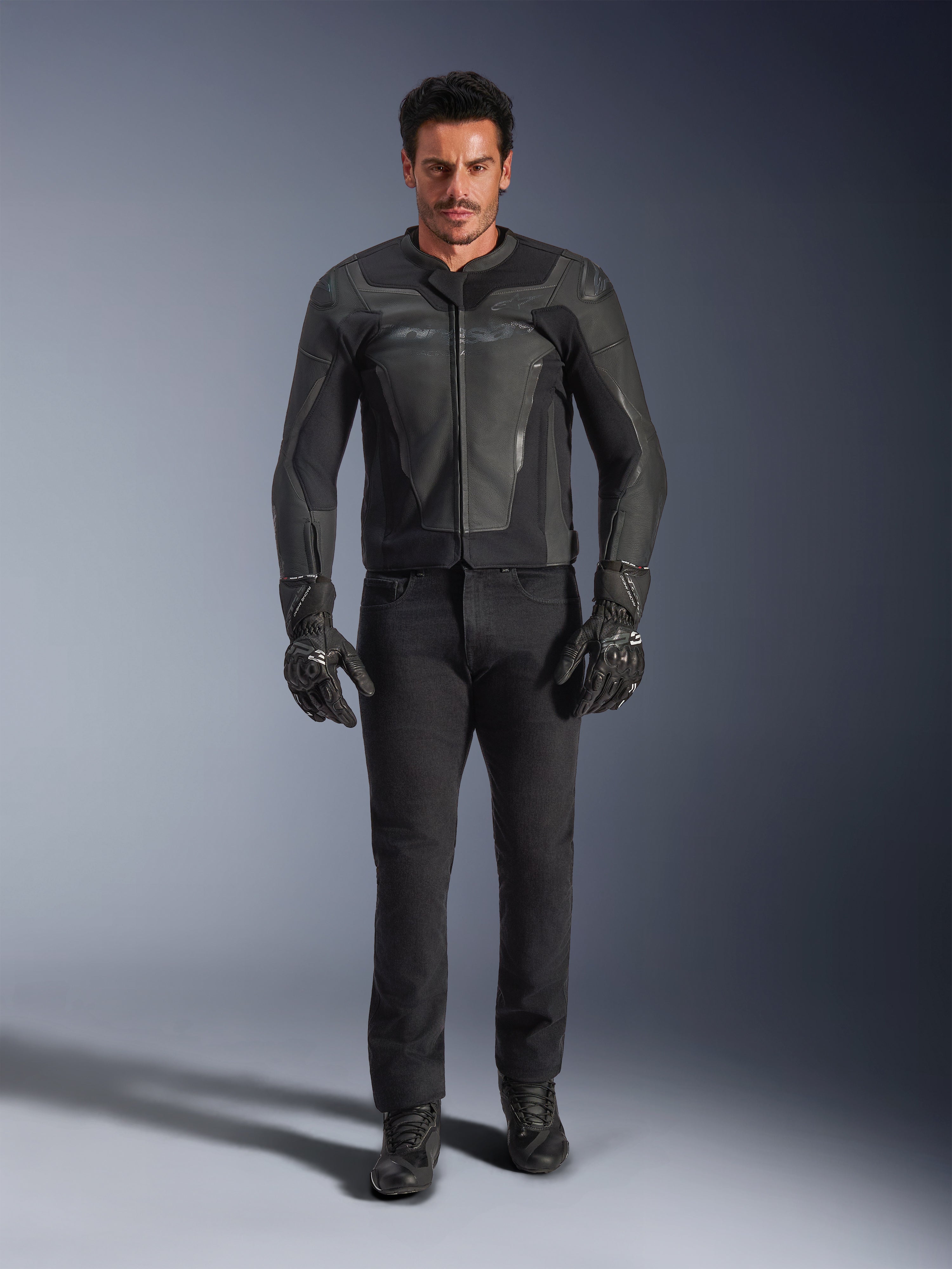 Veste en cuir Alpinestars GP Force V2 noire, portée par un motard avec un pantalon de moto, des gants et des bottes noirs, construction en cuir épurée de style sportif avec sliders d'épaules et protection intégrée.