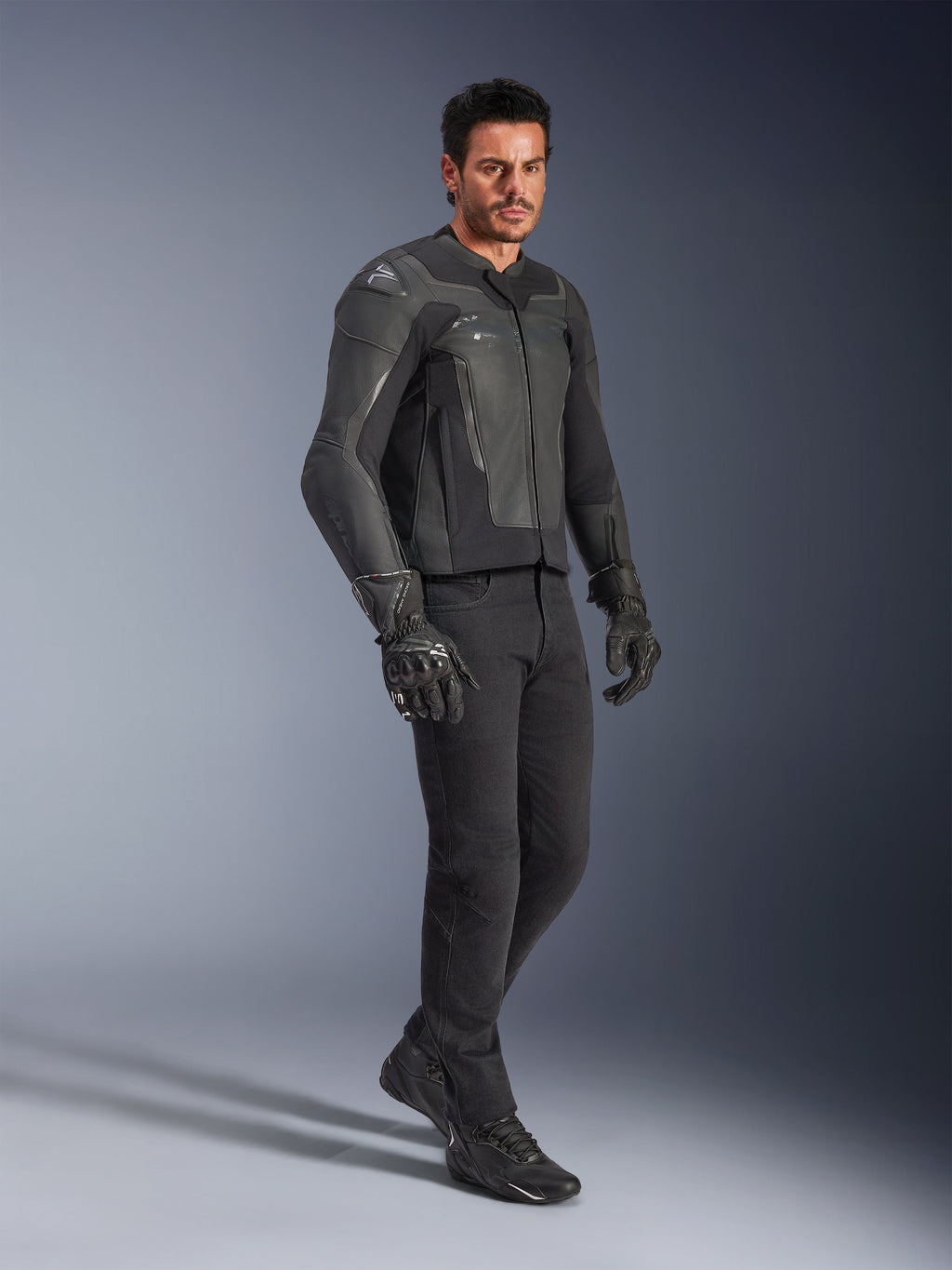 Homme portant une veste en cuir Alpinestars GP Force V2 noire, avec un pantalon noir et des gants de moto, photo de studio en pied, confection en cuir de bovin avec protections d'épaules et de coudes intégrées, équipement de route professionnel pour moto de sport