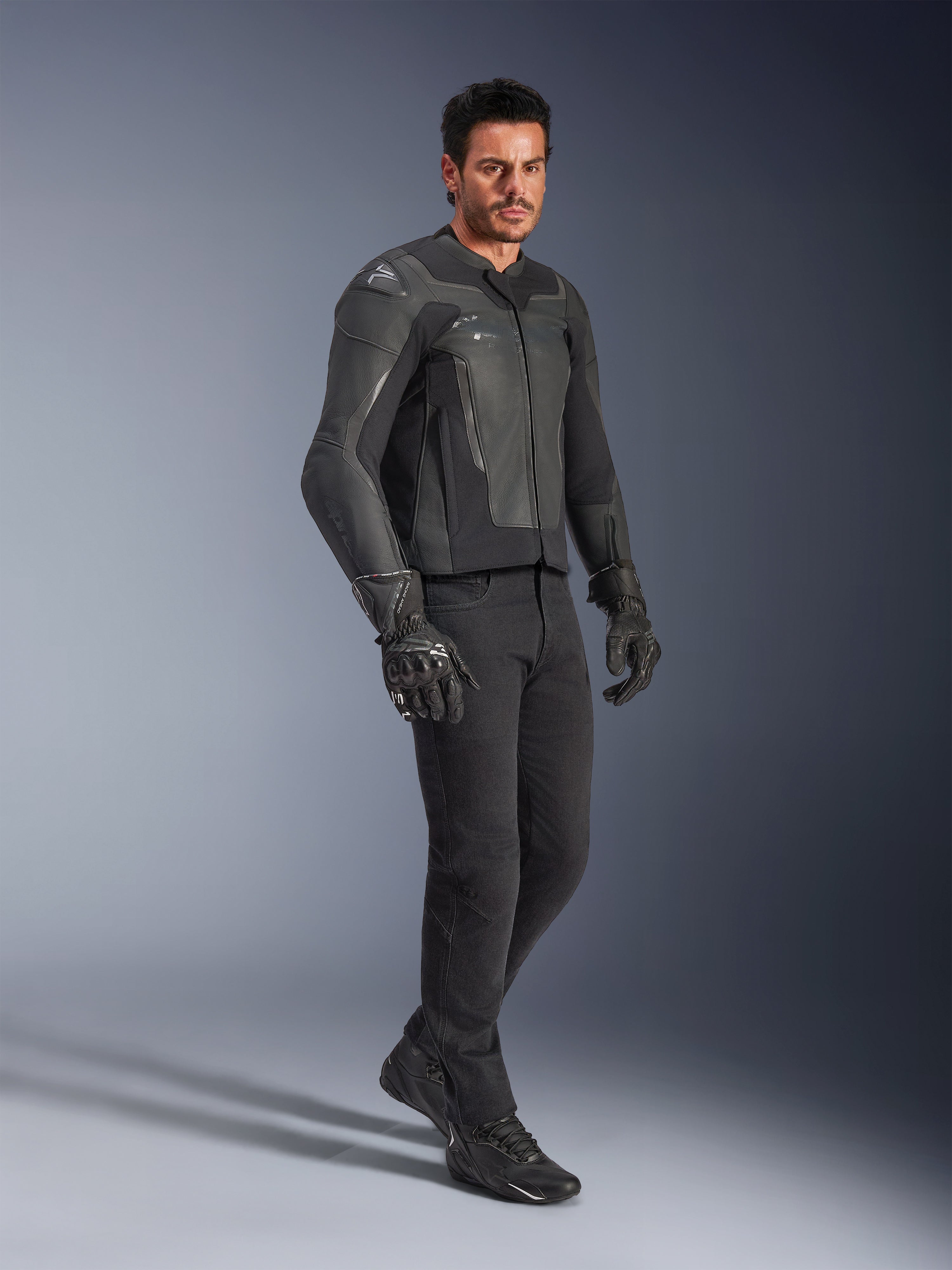 Homme portant une veste en cuir Alpinestars GP Force V2 noire, avec un pantalon noir et des gants de moto, photo de studio en pied, confection en cuir de bovin avec protections d'épaules et de coudes intégrées, équipement de route professionnel pour moto de sport