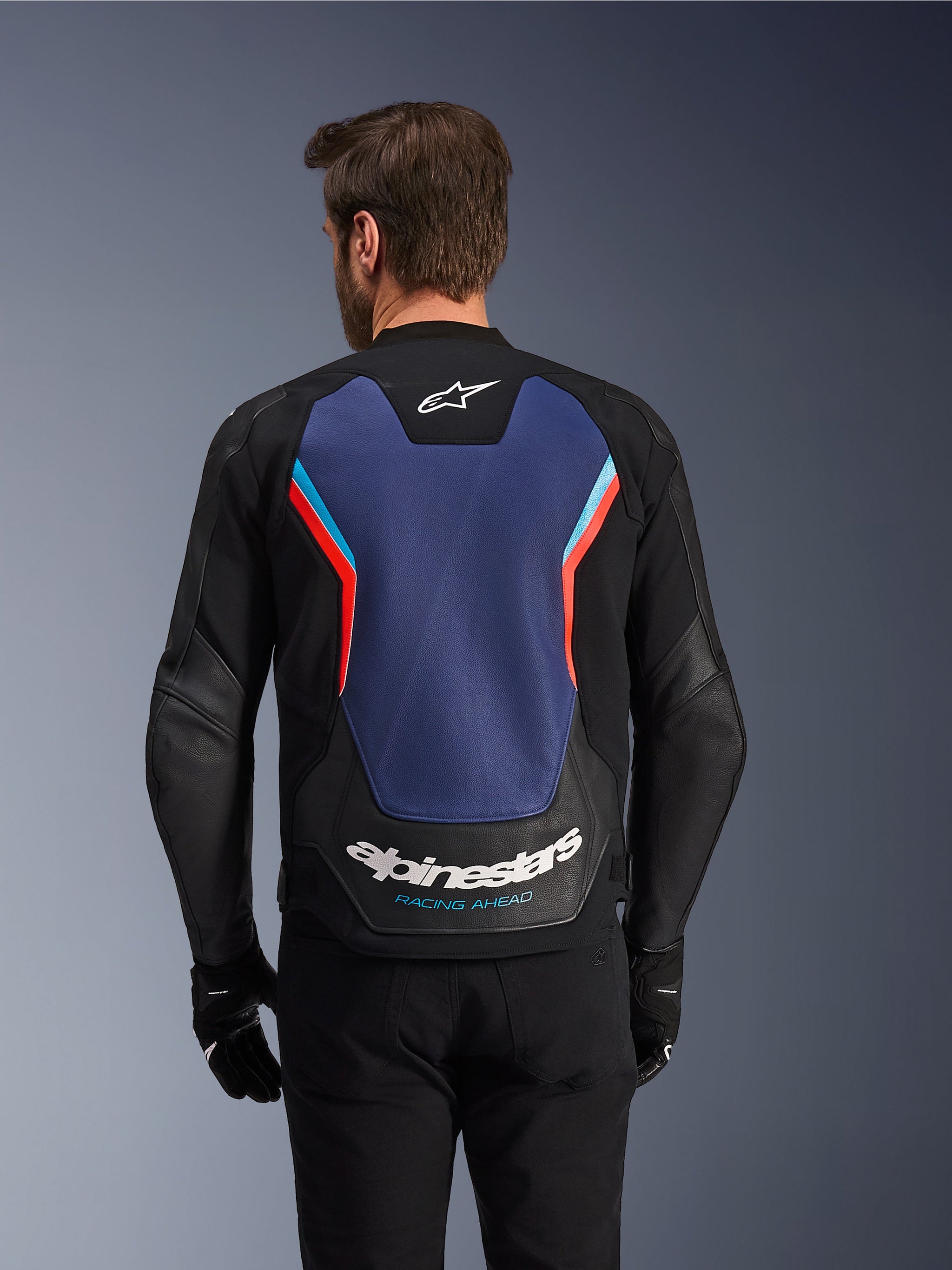Vue arrière du blouson en cuir Alpinestars GP Force V2 en noir, bleu et rouge fluo, présentant un large panneau dorsal bleu avec des touches de rouge fluo et de bleu clair, le logo Alpinestars blanc et l'inscription « Racing Ahead » dans le bas du dos, porté par un pilote avec des gants et un pantalon noirs.