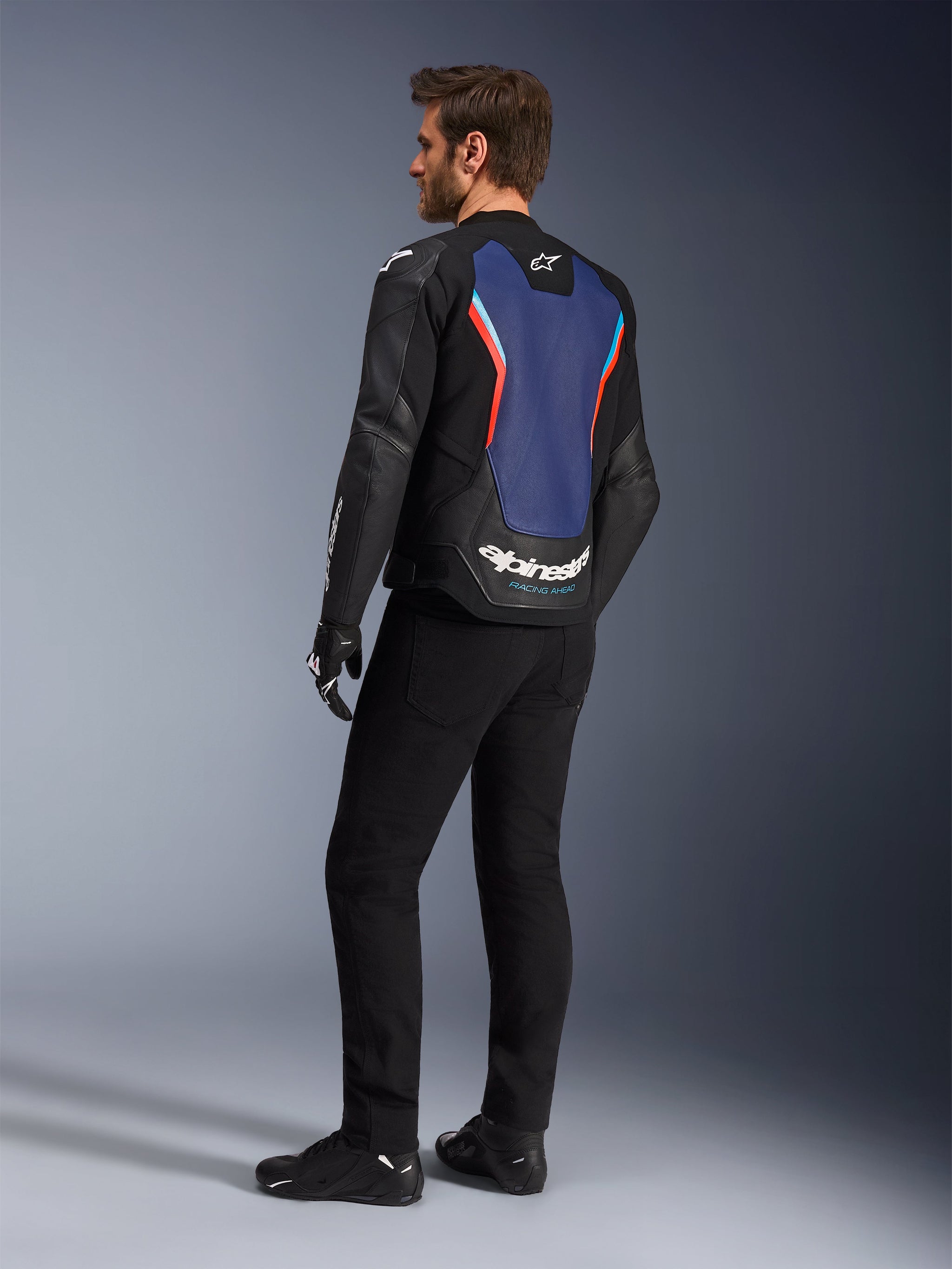 Vue de trois-quarts arrière d'un mannequin portant le blouson en cuir Alpinestars GP Force V2 en Noir Bleu Rouge Fluo, mettant en valeur le panneau arrière bleu avec des accents rouge fluorescent et des logos blancs, assorti à un pantalon noir et des gants de moto.