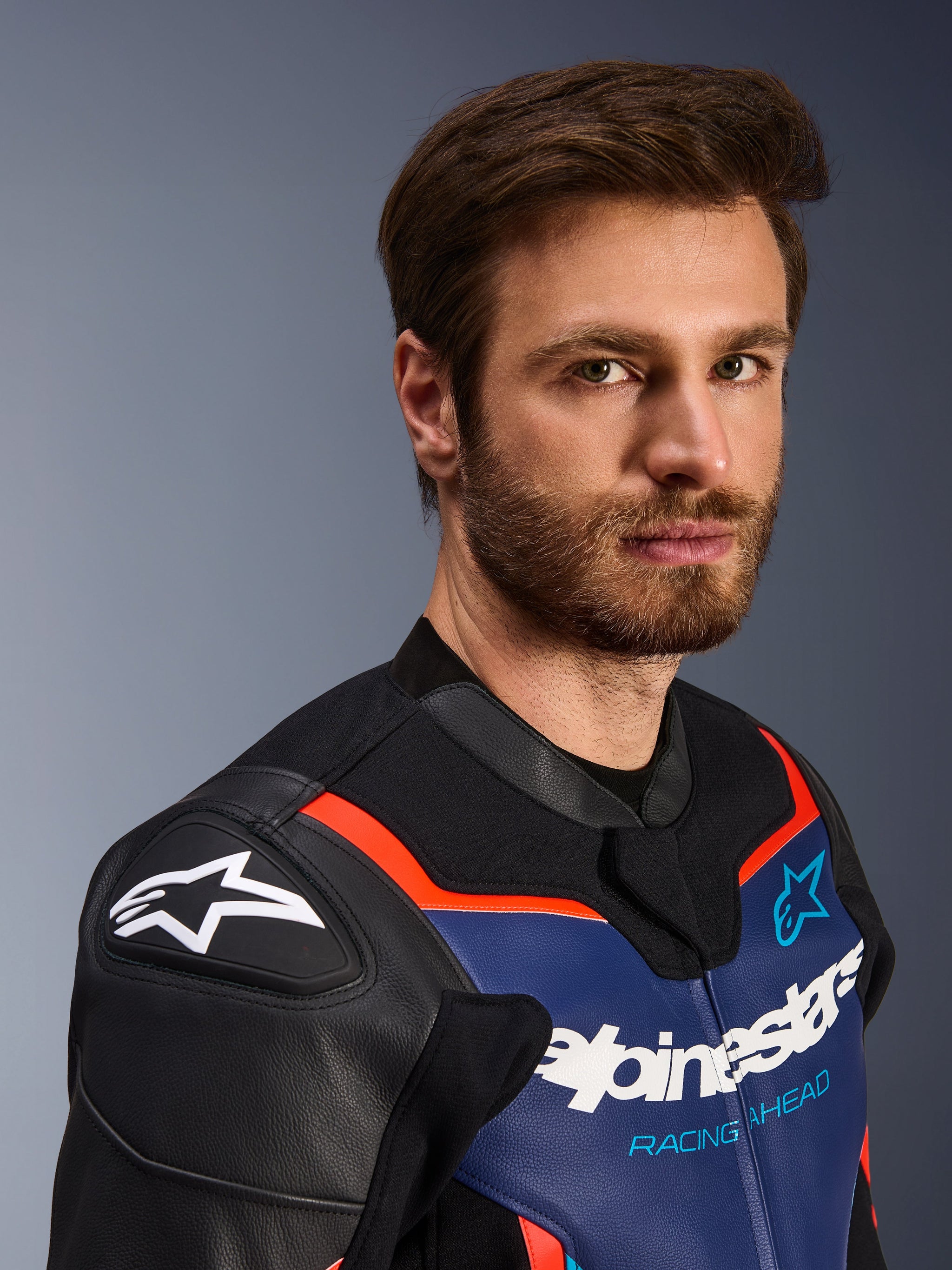 Blouson en cuir Alpinestars GP Force V2, noir, bleu et rouge fluo, portrait en gros plan d'un pilote masculin, arborant le logo Alpinestars blanc sur les protections d'épaules DFS noires, empiècement de poitrine en cuir bleu avec marquage blanc et texte 'Racing Ahead', accents rouge fluo, équipement de moto sportif haut de gamme.