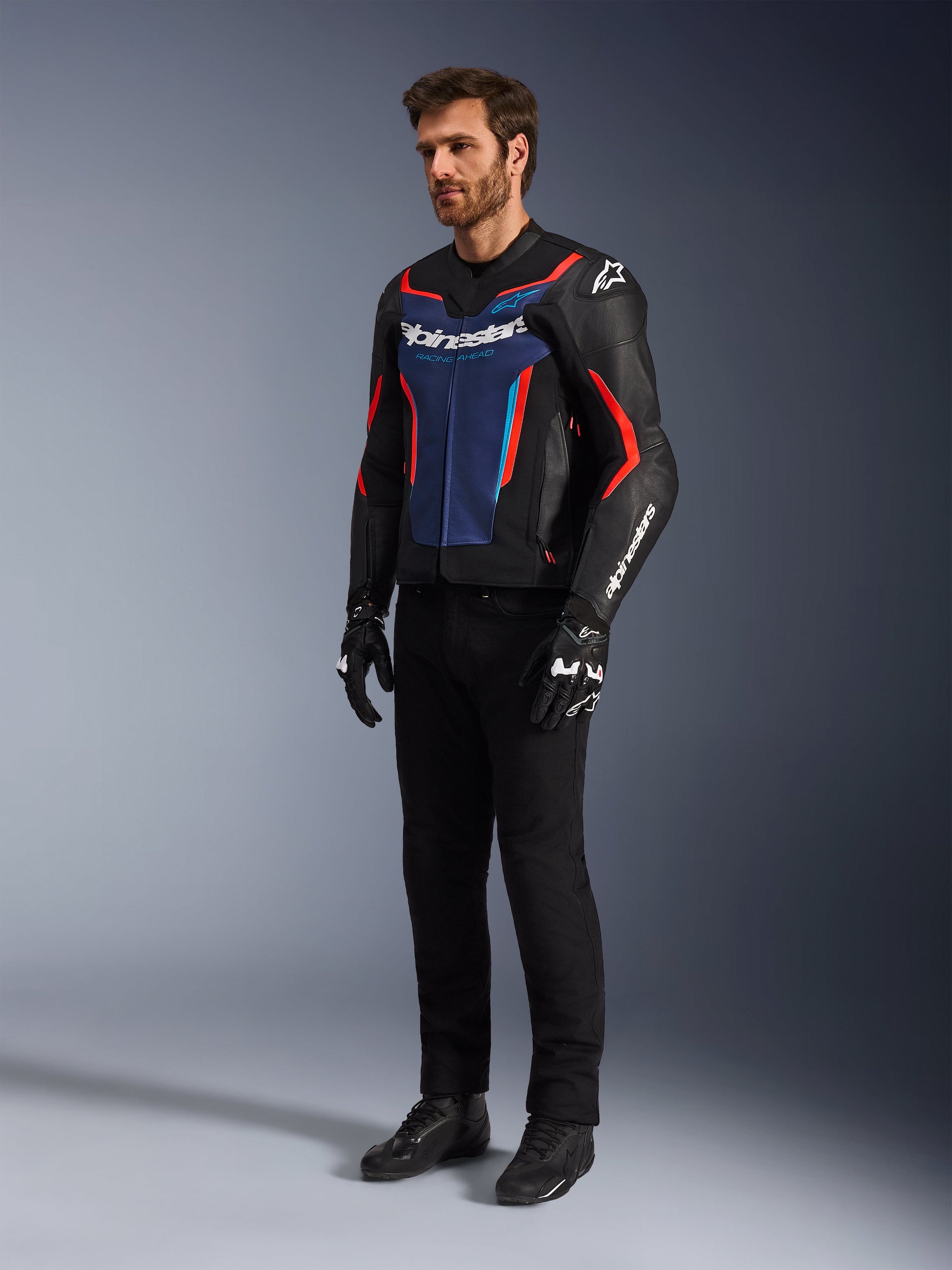 Modèle portant le blouson en cuir Alpinestars GP Force V2 en noir, bleu et rouge fluo, associé à un pantalon de moto technique noir, des gants noirs et des bottes, équipement de moto professionnel pour la conduite sportive