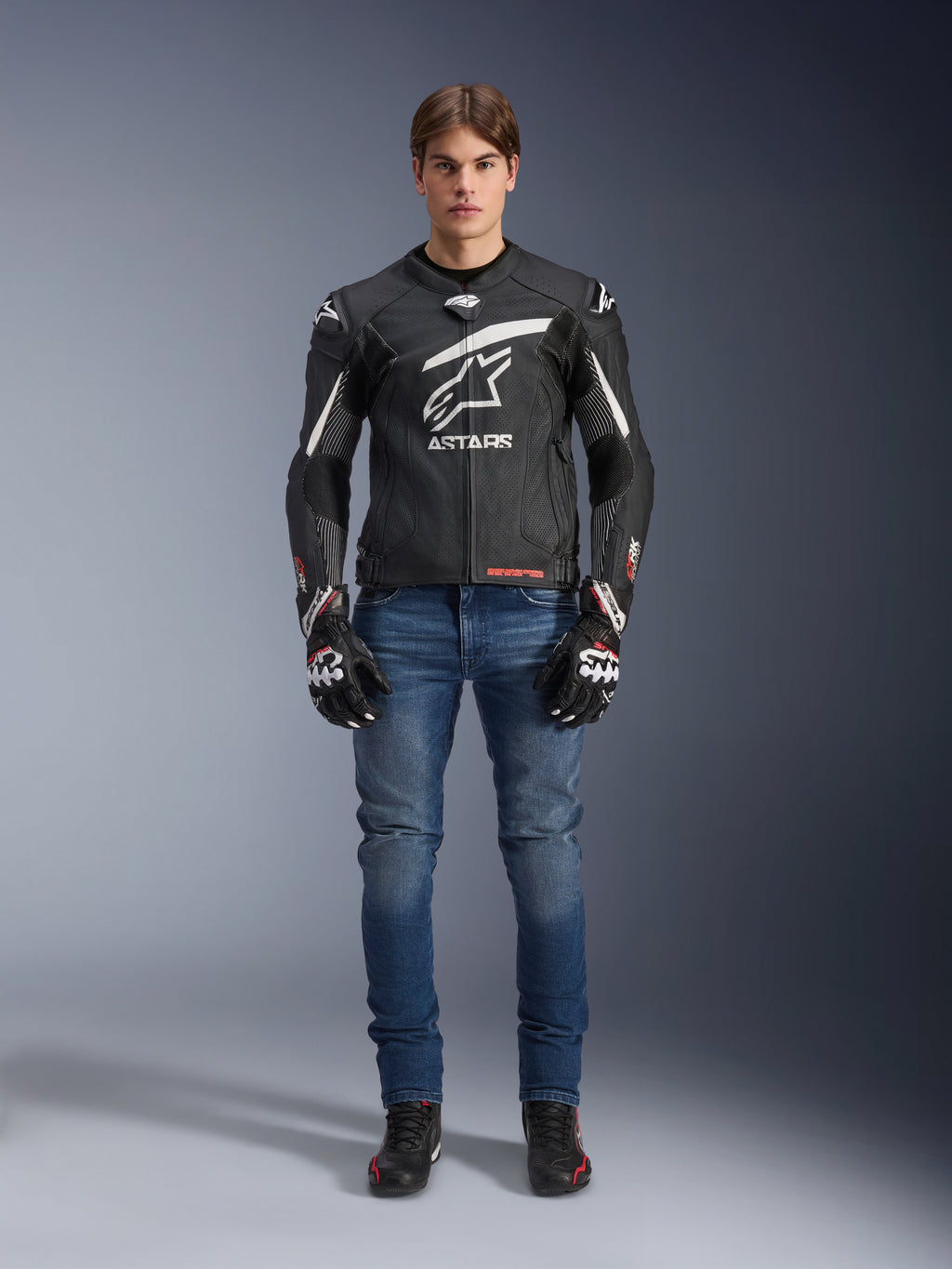 Male model wearing Alpinestars GP Plus R V4 Rideknit Leather Jacket in Black and White, styled with blue riding jeans, black motorcycle gloves, and black riding shoes, featuring a large white Astars logo on the chest, perforated leather panels, and white racing stripes on the sleeves, full-body standing view