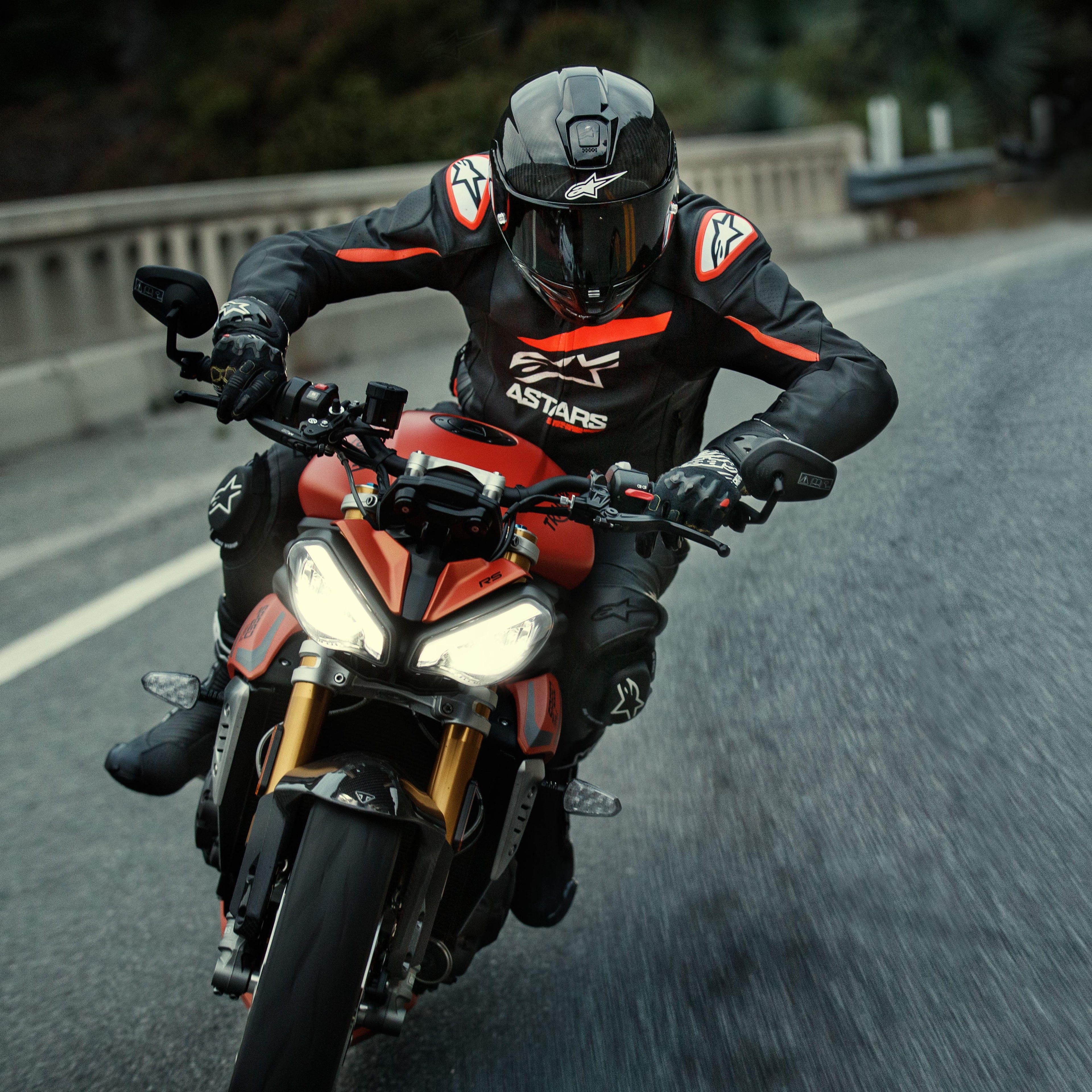 Motard sur une moto de sport rouge, veste en cuir Alpinestars GP Plus R V4 Rideknit, noir avec détails rouge fluo et blancs, logo Alpinestars blanc et texte 'ASTARS', casque et gants de course noirs, équipement de protection haute performance en action sur la route.