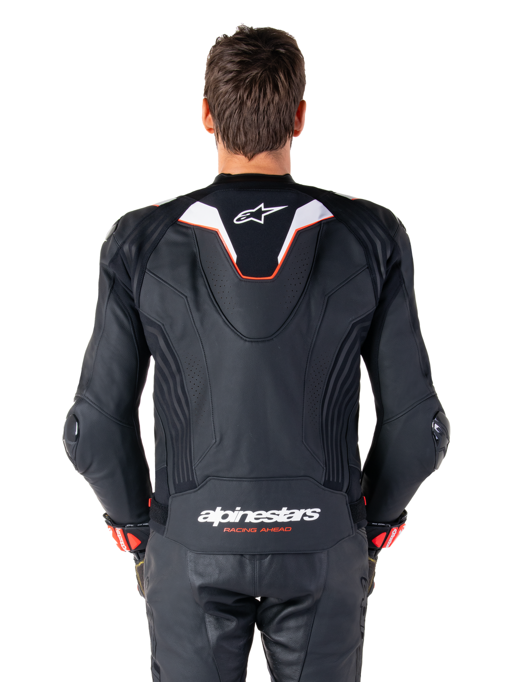 Vue de dos d'un pilote portant la veste en cuir Alpinestars Missile V3 Ignition, cuir noir avec accents blancs et rouges, dotée d'une bosse aérodynamique sport avec un logo blanc et le marquage 'alpinestars RACING AHEAD' dans le bas du dos, associée à un pantalon et des gants de course en cuir noir assortis.