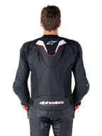 Vue de dos d'un pilote portant la veste en cuir Alpinestars Missile V3 Ignition, cuir noir avec accents blancs et rouges, dotée d'une bosse aérodynamique sport avec un logo blanc et le marquage 'alpinestars RACING AHEAD' dans le bas du dos, associée à un pantalon et des gants de course en cuir noir assortis.
