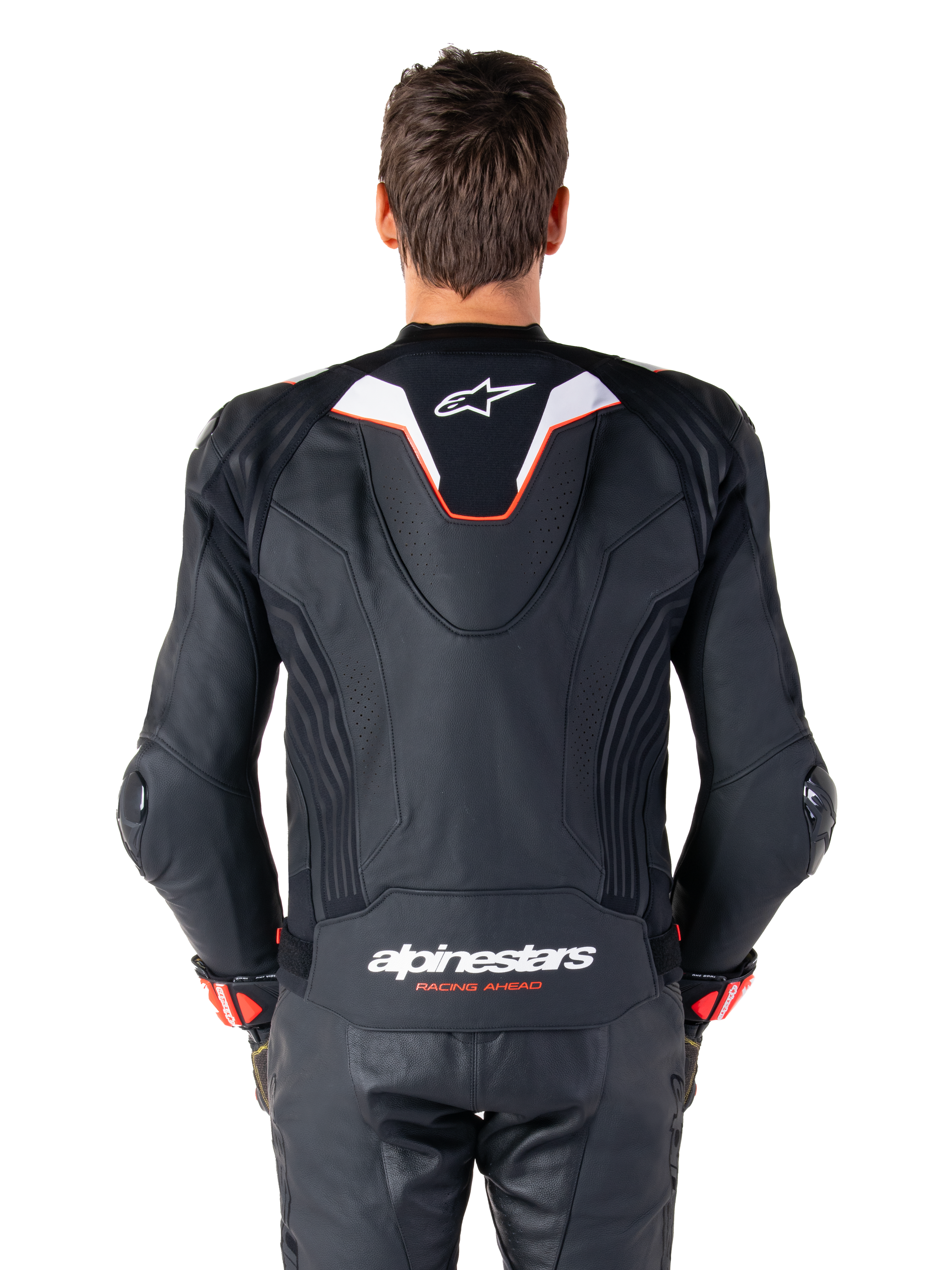 Vue de dos d'un pilote portant la veste en cuir Alpinestars Missile V3 Ignition, cuir noir avec accents blancs et rouges, dotée d'une bosse aérodynamique sport avec un logo blanc et le marquage 'alpinestars RACING AHEAD' dans le bas du dos, associée à un pantalon et des gants de course en cuir noir assortis.