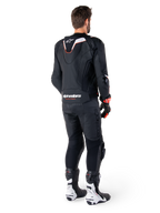 Vue arrière d'un pilote portant la veste en cuir Alpinestars Missile V3 Ignition noire, avec une bosse aérodynamique sport aux accents blancs et rouges, marquage Alpinestars au bas du dos, associée à un pantalon en cuir noir et des bottes de course noires et blanches, tenue de pilotage sportive professionnelle