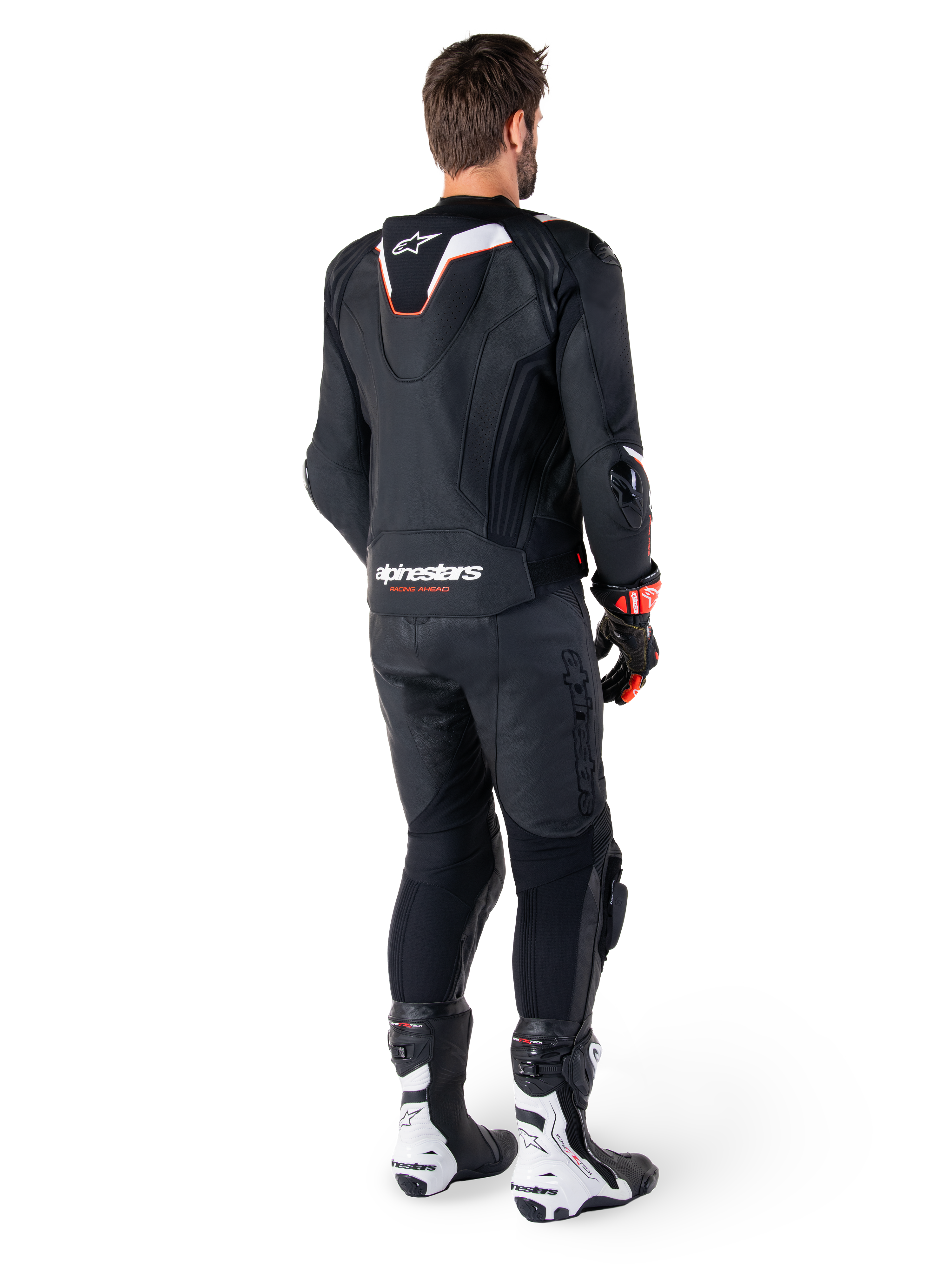 Vue arrière d'un pilote portant la veste en cuir Alpinestars Missile V3 Ignition noire, avec une bosse aérodynamique sport aux accents blancs et rouges, marquage Alpinestars au bas du dos, associée à un pantalon en cuir noir et des bottes de course noires et blanches, tenue de pilotage sportive professionnelle
