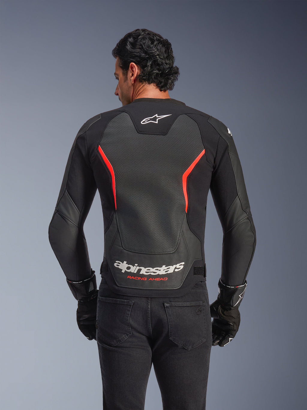 Blouson en cuir Alpinestars GP Force V2 Air, vue de dos, noir et rouge fluo, avec un large panneau dorsal en cuir perforé pour la ventilation, logo « Alpinestars Racing Ahead » dans le bas du dos, porté par un pilote avec des gants noirs, équipement de moto sportif haute performance