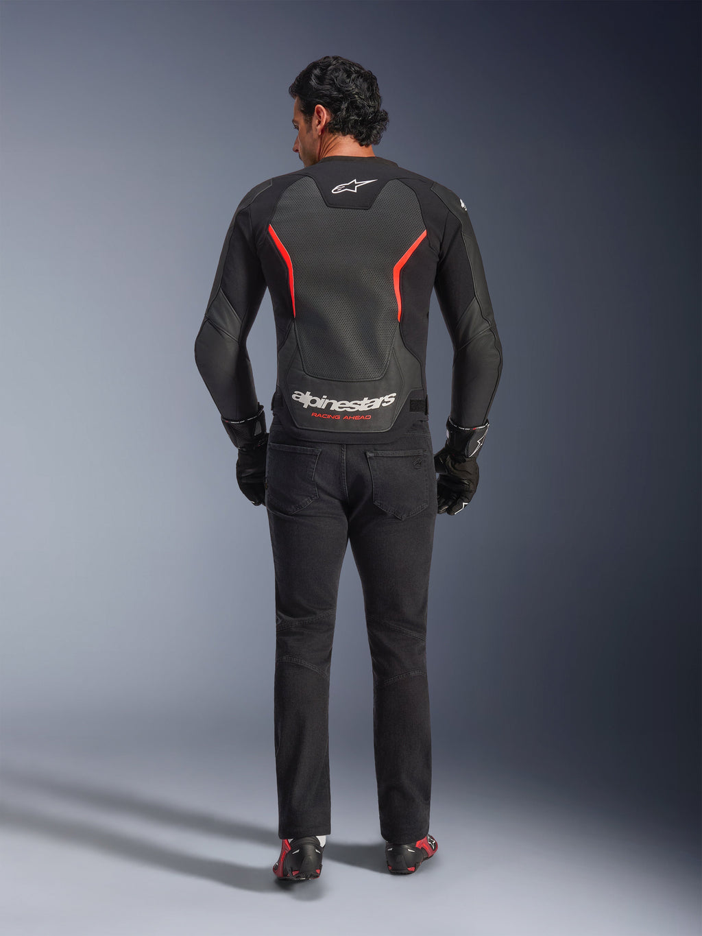 Blouson en cuir Alpinestars GP Force V2 Air en noir et rouge fluo, vue de dos sur mannequin, doté de panneaux dorsaux en cuir perforé, d'accents rouges et du marquage Alpinestars, porté avec un jean de moto noir et des gants pour un look de moto sportive.