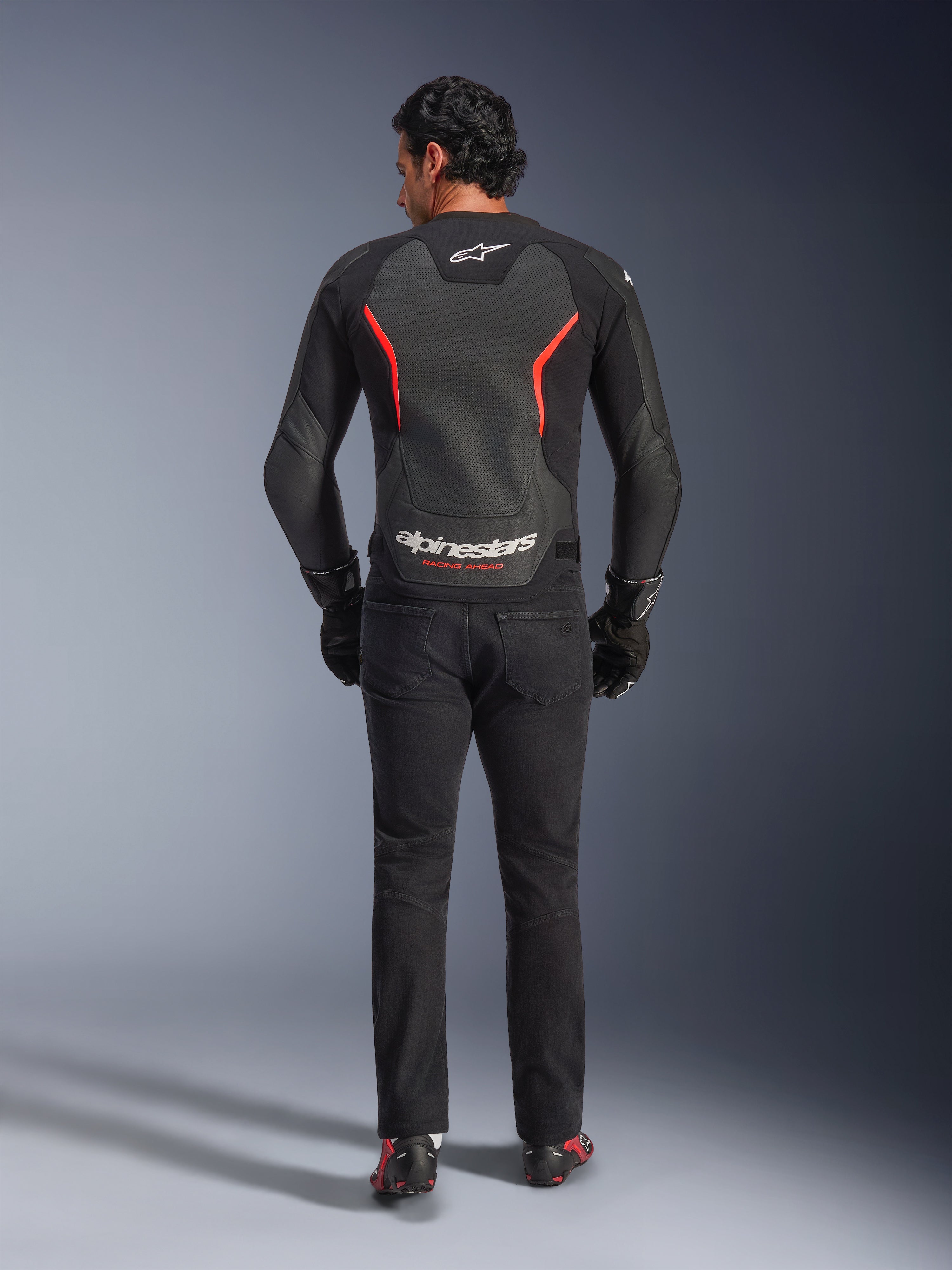 Blouson en cuir Alpinestars GP Force V2 Air en noir et rouge fluo, vue de dos sur mannequin, doté de panneaux dorsaux en cuir perforé, d'accents rouges et du marquage Alpinestars, porté avec un jean de moto noir et des gants pour un look de moto sportive.