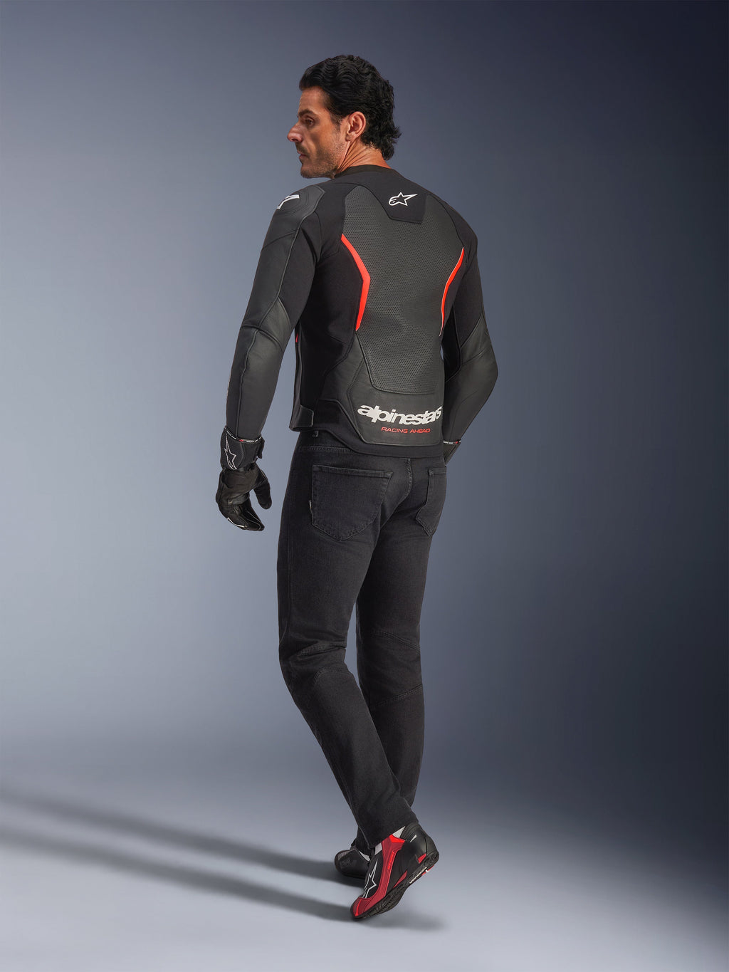 Modèle masculin vu de dos portant le blouson en cuir Alpinestars GP Force V2 Air en Noir Rouge Fluo, présentant de larges perforations dorsales et un marquage blanc, associé à un jean de moto noir, des gants et des bottes de moto noires et rouges en studio.