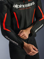Blouson en cuir Alpinestars GP Force V2 Air en Noir et Rouge Fluo, gros plan montrant la construction en cuir perforé, logos blancs sur la poitrine et les manches, et un pilote ajustant la fermeture éclair de l'avant-bras pour un ajustement sur mesure.