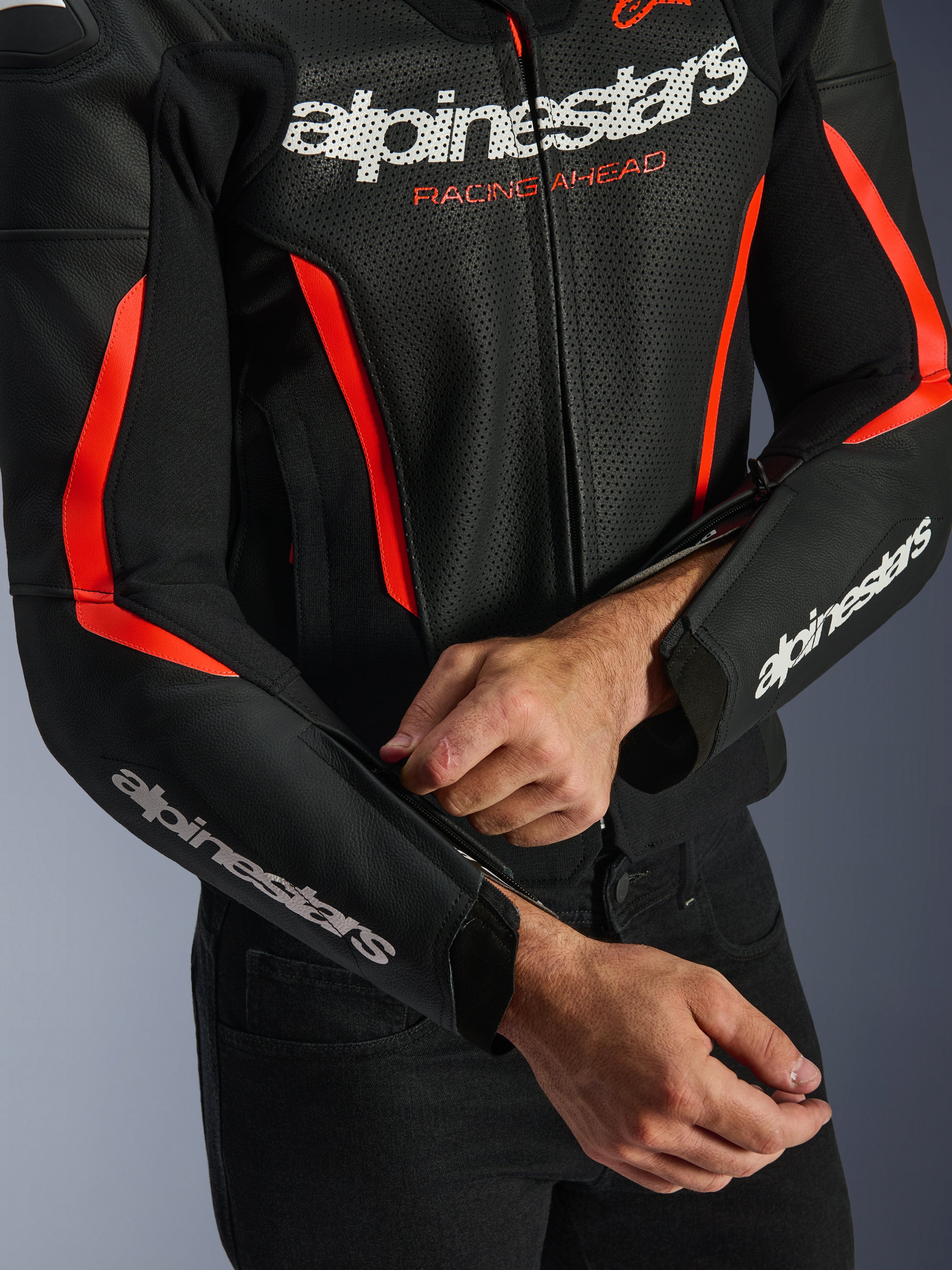 Blouson en cuir Alpinestars GP Force V2 Air en Noir et Rouge Fluo, gros plan montrant la construction en cuir perforé, logos blancs sur la poitrine et les manches, et un pilote ajustant la fermeture éclair de l'avant-bras pour un ajustement sur mesure.