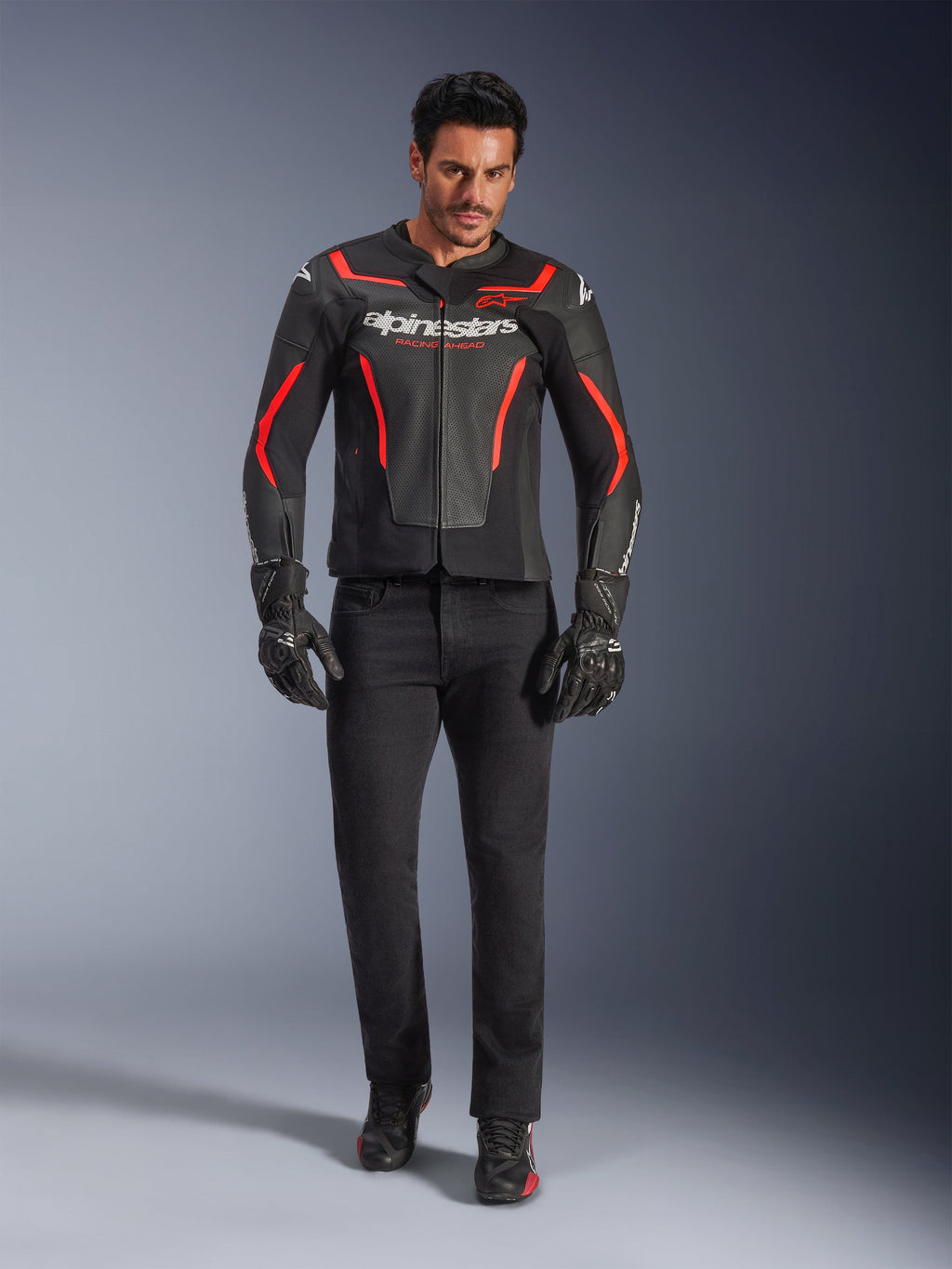 Pilote portant le blouson en cuir Alpinestars GP Force V2 Air, noir et rouge fluo, associé à un jean de moto noir, des gants et des chaussures, avec une construction en cuir perforé, des logos blancs et des accents rouges élégants pour un look sportif